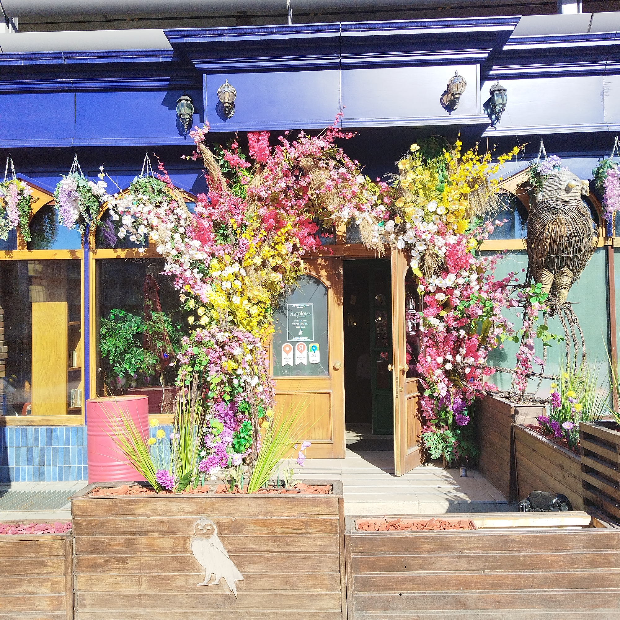 The image displays the vibrant and whimsically decorated facade of a storefront, likely a cafe or restaurant, located in Yekaterinburg, Russia. The building features a deep blue upper facade with ornate trim and vintage-style wall lamps, set under a bright, sunny sky that casts distinct shadows.
The main entrance, consisting of open double wooden doors, is framed by an elaborate archway of artificial flowers in a riot of pink, yellow, white, and purple hues, intertwined with dried grasses. Large glass windows flank the doorway, reflecting the outdoor light and offering glimpses of interior greenery. On the glass of the right door, a clear sign displays the name "ПЛАТФОРМА" (PLATFORMA) and operating hours "12:00 - 22:00," along with three badges that appear to be awards or ratings from local review platforms.
Along the front, several wooden planters are filled with green plants and more colorful flowers. A decorative wooden cutout of an owl is attached to one planter, while a much larger, intricately woven wicker owl sculpture stands prominently to the right of the entrance. Other notable details include a bright pink barrel positioned next to a planter on the left and a base of light blue tiles beneath the windows. No people are visible, but the open doors and festive decor create an inviting and cheerful atmosphere, typical of a lively urban establishment.