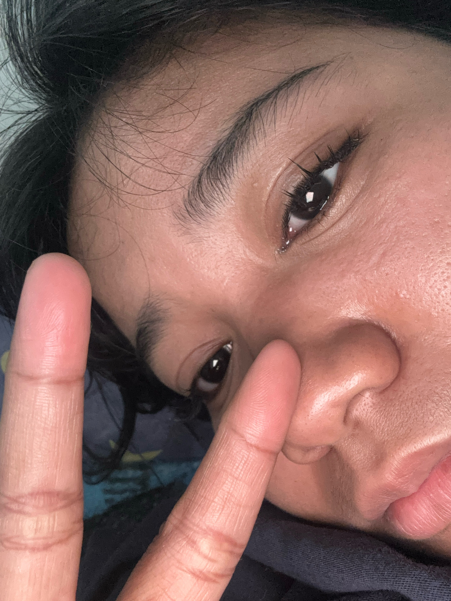The image shows a close-up, angled selfie of a person's face, specifically focusing on their left eye, nose, and mouth. The person is holding up two fingers, forming a peace sign, partially obscuring their face. Their skin appears to have visible pores and some texture. The background is dark, with hints of blue and yellow fabric visible. The lighting is artificial and appears to be from above, creating some shadows on the face. The overall impression is that of a casual, personal photograph. This image was taken at a residence in North Jakarta, Indonesia.