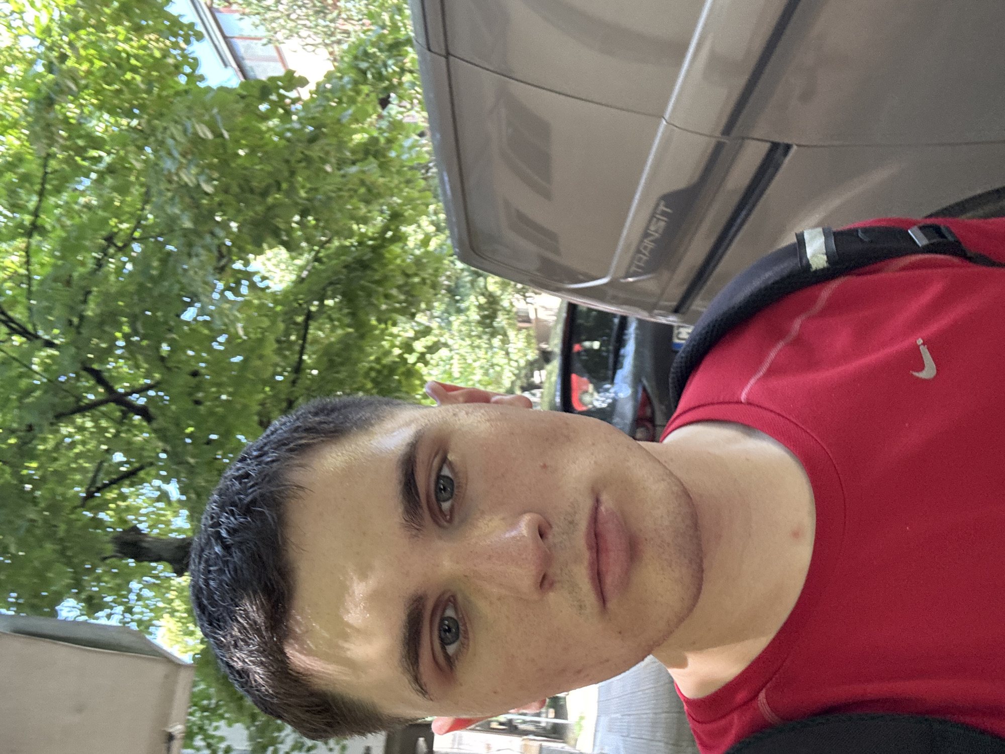 In Stara Zagora, Bulgaria, a young man with short, dark hair and fair skin takes a selfie outdoors. He has light-colored eyes, and his face is slightly flushed with some freckles. He is wearing a red t-shirt with a Nike logo and a black backpack. Behind him, a beige van with "TRANSIT" written on its side is parked, and a tree with lush green leaves provides shade. The background also shows a building and another car. The lighting suggests it is daytime, possibly late morning or early afternoon, as indicated by the dappled sunlight on the man's face.