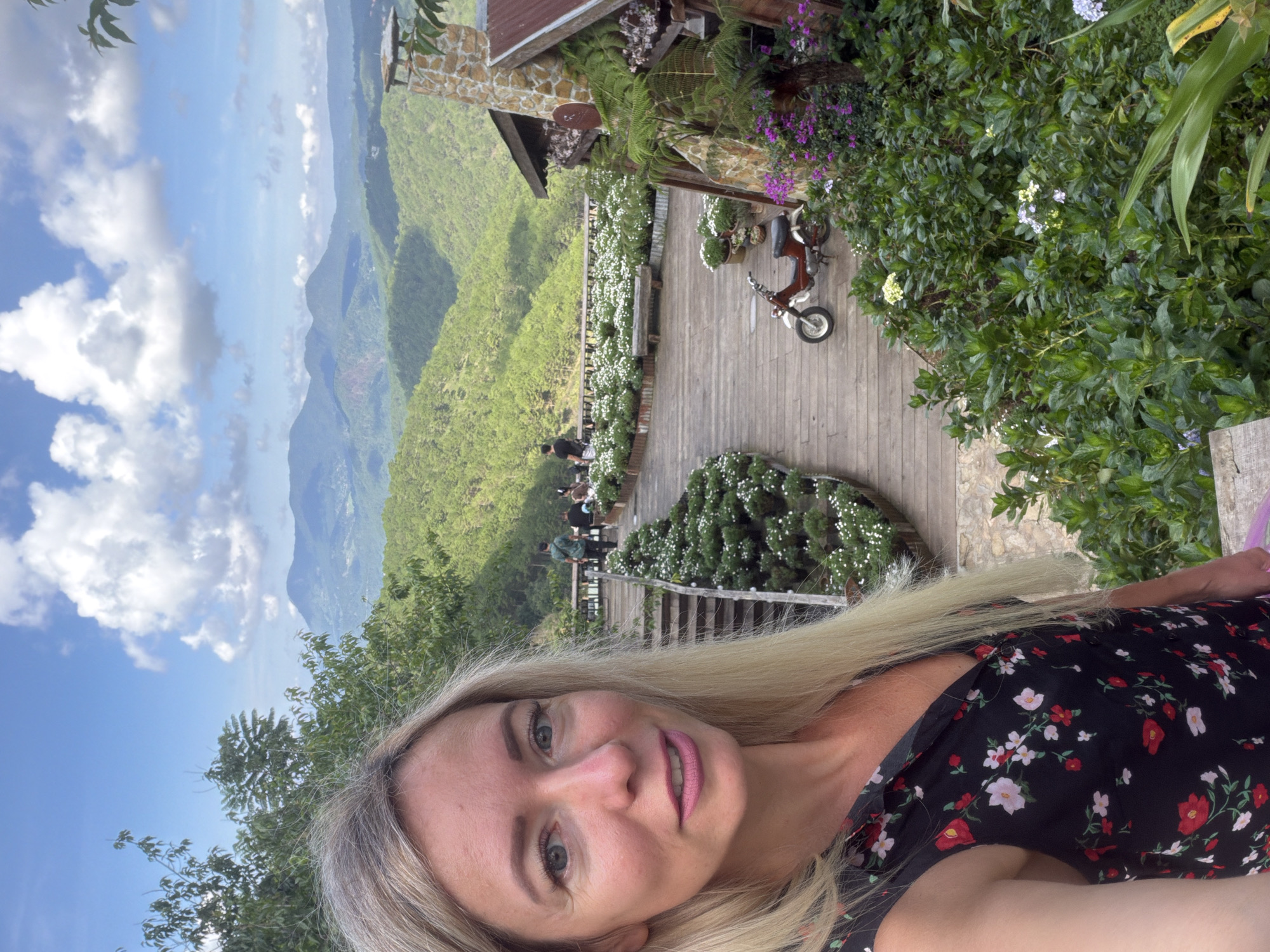 The image depicts a scenic outdoor setting with a woman in the foreground. She has blonde hair and is wearing a black dress with a floral pattern. She is facing the camera and smiling. Behind her, a wooden pathway winds through lush greenery and leads to a building with a stone chimney. The pathway is lined with numerous small white flowers. In the background, rolling green hills covered with trees stretch out under a bright blue sky with scattered white clouds. Several people are visible walking along the pathway in the middle ground. A small red and white motorcycle is parked on the pathway. The overall impression is a tranquil, natural landscape with a touch of rustic charm. This location is identified as being in Xuân Thành, Vietnam.