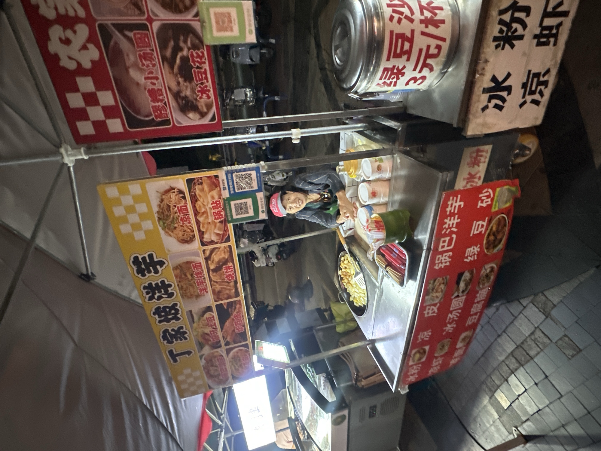 An outdoor food stall is set up under a large canopy, likely at night, as suggested by the artificial lighting and the presence of streetlights in the background. The stall displays a variety of food items, with prominent signage in Chinese characters. One sign, in yellow and white, features pictures of different dishes, including noodles and dumplings. Another sign, in red and white, also displays food images, with text that appears to be menu items.
A woman, wearing a red cap and a dark jacket, is the vendor. She is smiling and facing the camera, positioned behind a stainless steel food preparation counter. On the counter, there are several containers, some of which appear to hold condiments or ingredients, and a metal pot containing fried food, possibly potatoes or dough balls, being handled with a spatula. Red chopsticks are neatly arranged in a green holder. To the right of the counter, a large silver tin with red Chinese lettering, which reads "Green Bean Soup 3 Yuan/Cup", indicates a specific menu item. In front of the stall, a sign on a wooden plank displays "Ice Jelly Powder" in Chinese.
The ground is paved with cobblestones. In the background, several motorcycles are parked, suggesting this might be a busy street or market area. The overall scene depicts a typical street food vendor in what appears to be a lively urban environment. The location of the Chongqing High-tech Zone, China, is provided as context for this scene.