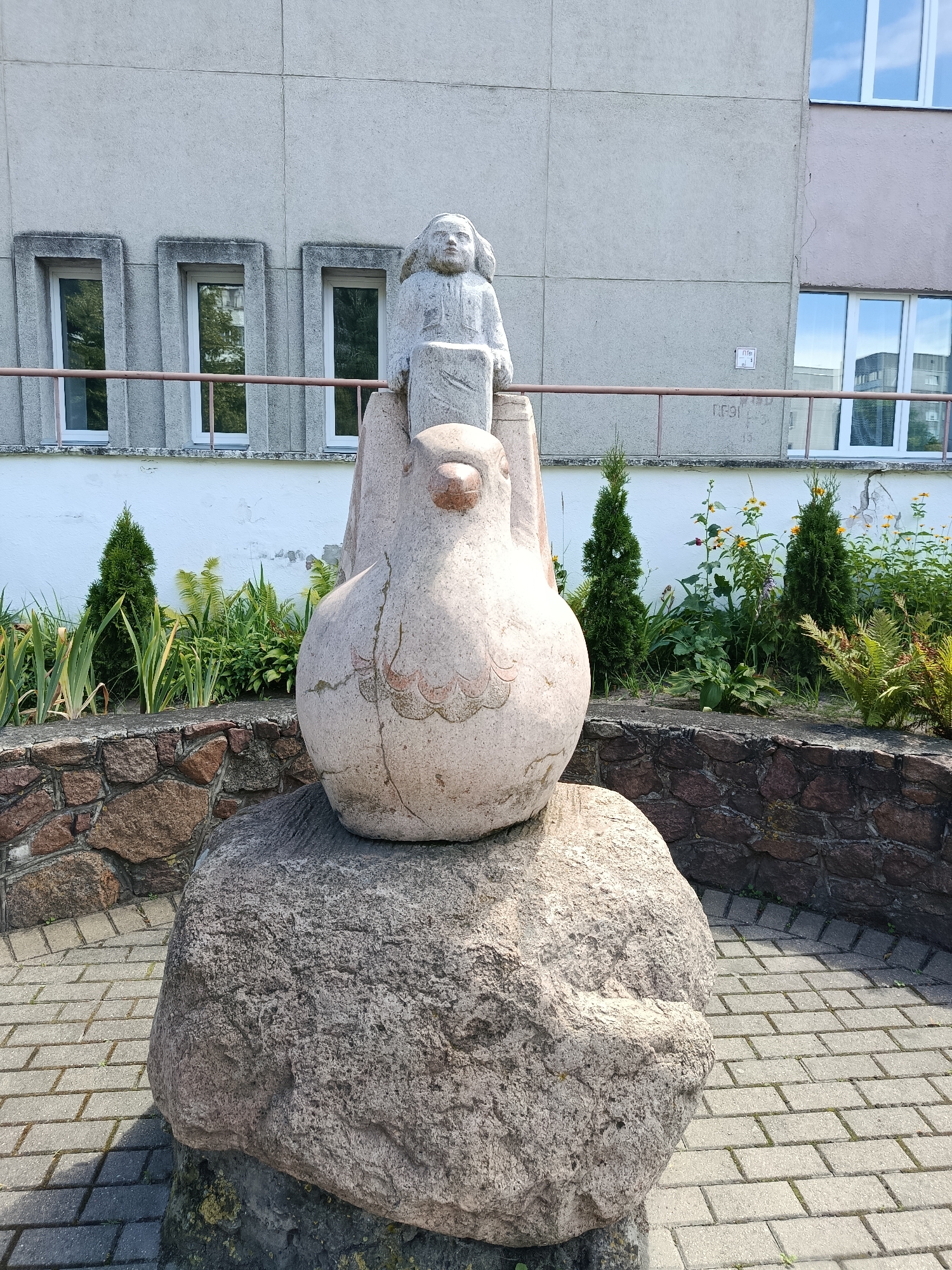 The image shows a stone sculpture of a bird with a child sitting on its back, located in the city of Svietlahorsk, Belarus. The sculpture is positioned on a large, rough-hewn rock. Behind the sculpture, a building with a light-colored facade and several rectangular windows can be seen. A metal railing runs along the base of the building. In front of the building and surrounding the sculpture, there is a garden area with greenery, including bushes and flowers. The ground around the sculpture is paved with light-colored rectangular stones. The lighting suggests it is a daytime scene with sunlight.