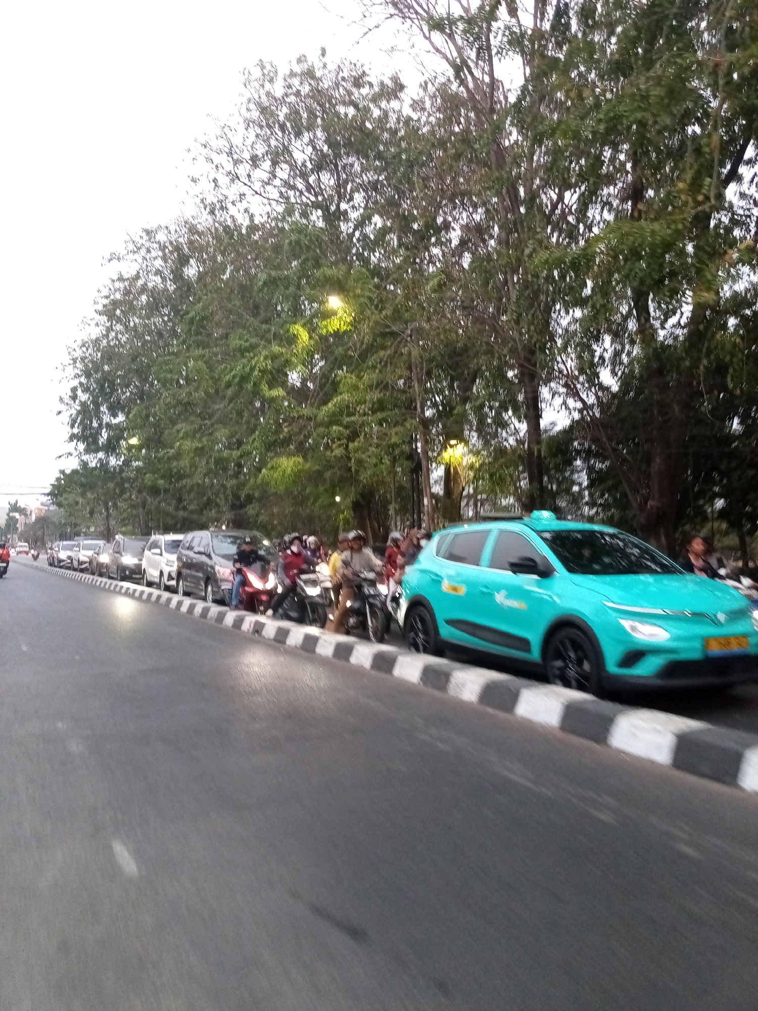 A turquoise colored taxi is parked alongside other vehicles on a street lined with trees. The taxi is a modern electric vehicle, with sleek headlights and black wheels. It has the taxi company's logo and text on its side. Several other cars and motorcycles are also parked on the street. Some of the motorcycles have riders on them, suggesting that they are either waiting or have just stopped. People are visible on the motorcycles and behind the parked cars, but their faces are not clearly discernible. The street is paved, and there is a raised curb with a black and white striped pattern separating the road from the sidewalk area. The background is filled with lush green trees, indicating an outdoor urban environment. The sky appears overcast. The scene suggests a busy street with traffic congestion, possibly in a residential or commercial area within the city of Bekasi, Indonesia.