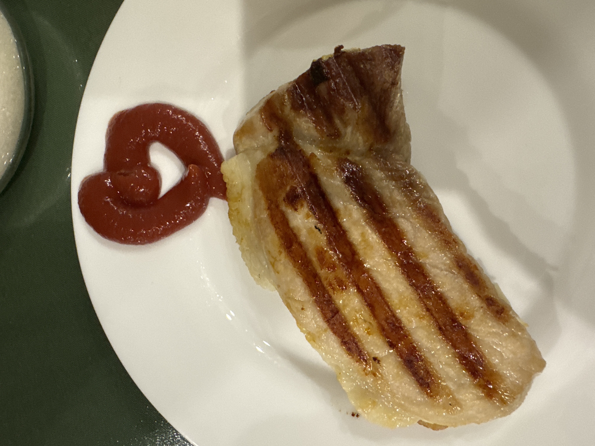 A grilled piece of meat, displaying prominent grill marks, is placed on a white plate next to a dollop of reddish-brown sauce. The plate is situated on a green surface. The surrounding environment is not identifiable, preventing the verification of its location in Votkinsk, Russia. No people or specific actions are visible in the image.