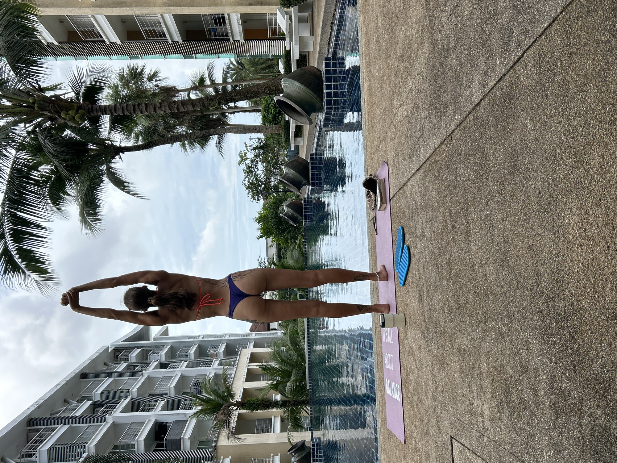 A full-body, rear-facing view shows a tanned woman in a bikini standing on a yoga mat next to a swimming pool. The woman has her arms raised above her head, hands clasped. She has a ponytail and tattoos on her back and legs. The yoga mat is pink and has text printed on it. To the right of the mat are two blue acupressure mats and a white water bottle. The swimming pool is long and rectangular, with a dark blue tiled edge. Decorative large, dark green pots are placed on pedestals along the pool's edge. In the background, there are modern apartment buildings with balconies and palm trees. The sky is overcast. The location is identified as Kathu, Thailand.