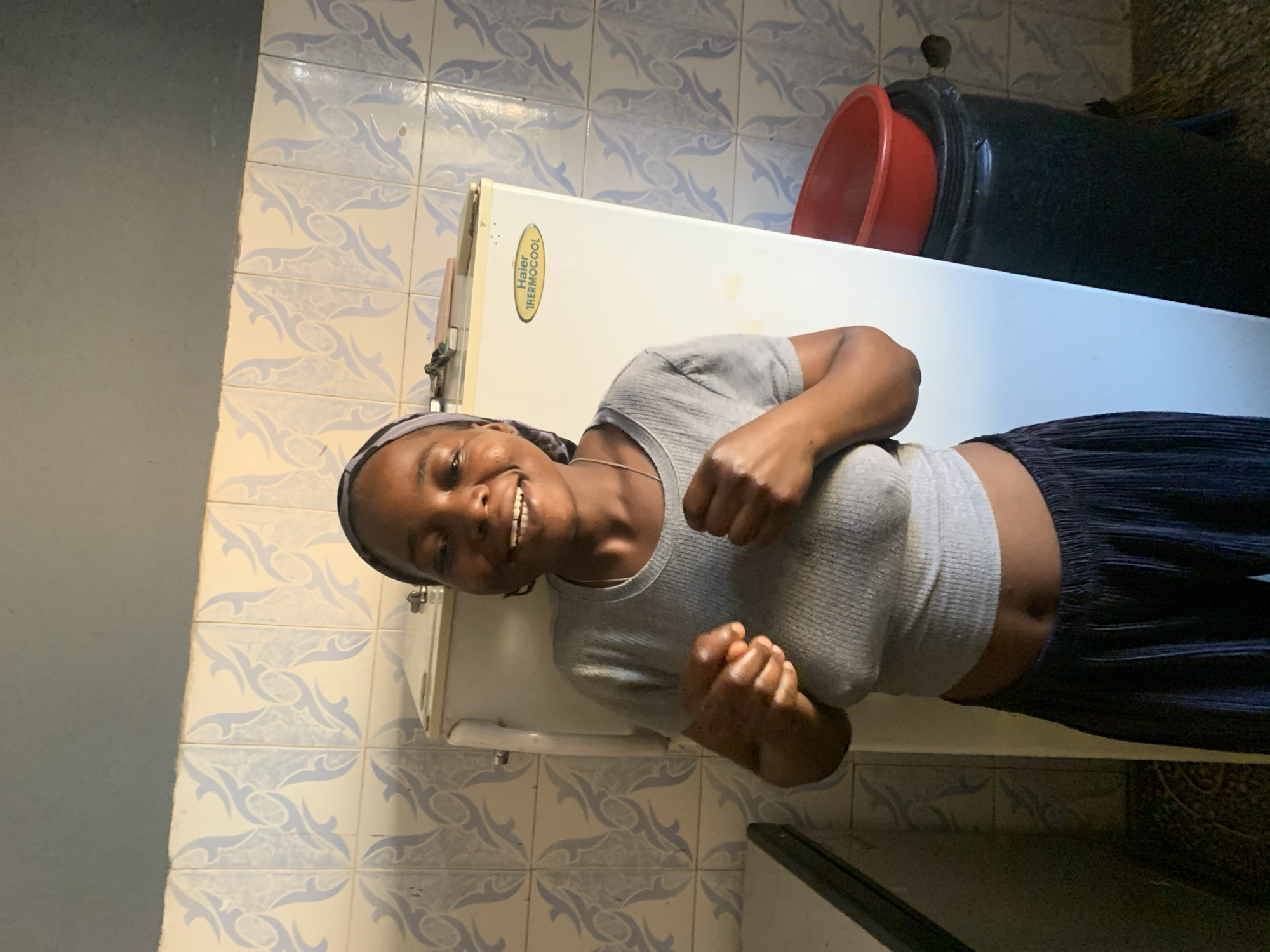 A young woman with dark skin and short dark hair, wearing a grey t-shirt and dark blue pleated pants, stands in front of a white refrigerator in a room with light blue and white patterned tiles on the wall. She is smiling and holding her fists up in front of her chest. The refrigerator has a yellow oval logo that reads "Haier Thermocool." To the right of the refrigerator, a black trash can is visible with a red plastic bowl on top. The floor is tiled with a dark pattern. The scene appears to be in a domestic setting, possibly a kitchen, in Jos, Nigeria.