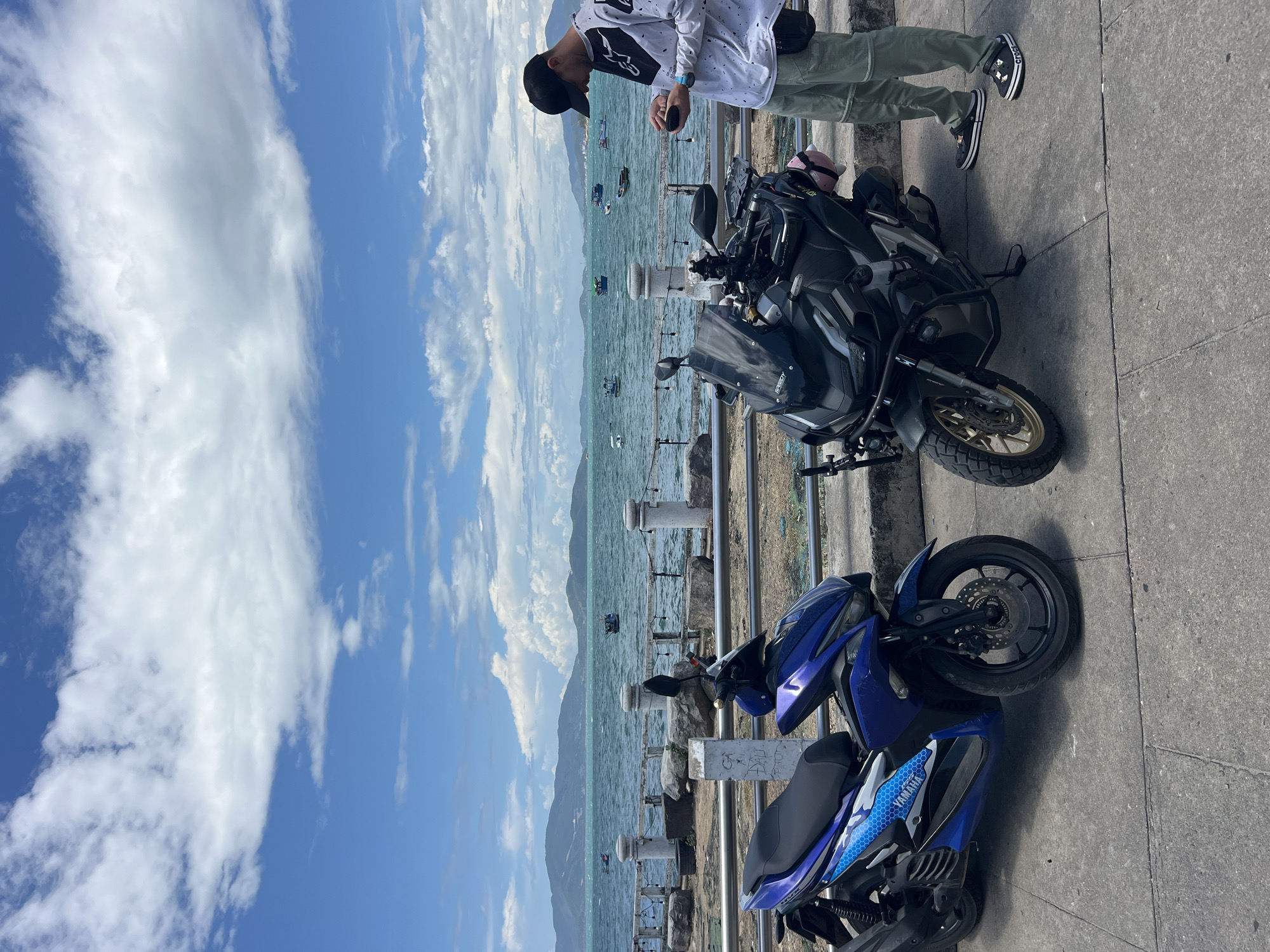 The image shows two motorcycles parked on a concrete walkway with the ocean in the background, likely in Bac Nha Trang Ward, Vietnam, indicated by the location mentioned. The motorcycle closer to the viewer is blue and has "Yamaha" branding. The other motorcycle is black and larger, appearing to be an adventure-touring bike. A person wearing a white patterned shirt, light green pants, and black shoes is standing next to the bikes, looking down at something in their hands. A low fence separates the walkway from the water. The sky is blue with scattered clouds. Several boats are visible on the water in the distance.