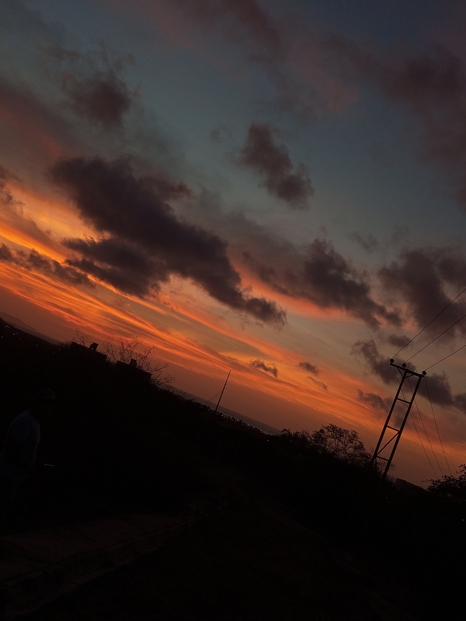 A long-angle shot captures a vivid sunset with orange and pink hues streaking across the sky, interspersed with dark, grey clouds. In the foreground, a silhouette of dark ground covers the lower portion of the frame, punctuated by sparse vegetation and what appears to be a power pole with wires extending outwards. The horizon shows a hint of urban development, suggesting the edge of the city. The location is Juan Griego, Venezuela. A figure can be seen to the far left of the foreground.
