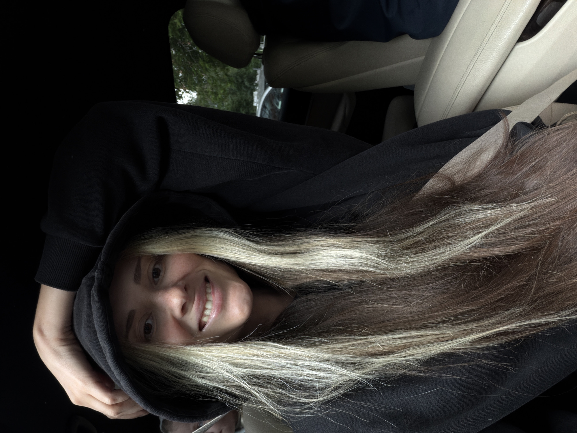 A fair-skinned female with long blonde and light brown hair is pictured wearing a black hooded sweatshirt inside a vehicle. The interior of the vehicle is visible, with beige leather seats and a seatbelt. A glimpse of trees can be seen through the car window. The image does not provide enough information to determine the exact location within the city of Khimki, Russia.