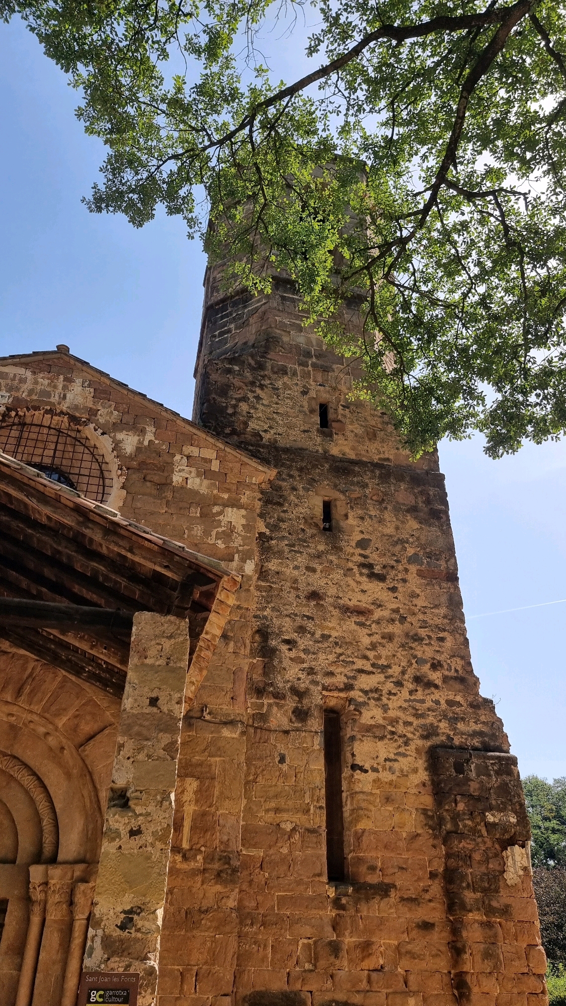This is a low-angle, eye-level, medium shot photograph of the Church of Sant Joan les Fonts, located in Sant Joan les Fonts, Spain. The main subject is the Romanesque bell tower, constructed from rough-hewn, light brown and tan stone blocks. The tower is tall and rectangular, with several small, narrow windows visible on its facade. To the left of the tower, a section of the church's facade is visible, also built from similar stone. This section features a rounded archway with intricate carvings and a large, circular window with a dark grille. A wooden roof structure juts out from the church, supported by stone pillars. Overhanging the top of the image, from the upper left to the upper right, are the branches and green leaves of a large tree, dappled with sunlight. The sky is a clear, pale blue. In the lower left corner, a small white sign with black text reads "Sant Joan les Fonts" and includes the logo "gc" and "garrotxa . cultur". The overall scene conveys a sense of historical architecture bathed in natural light.