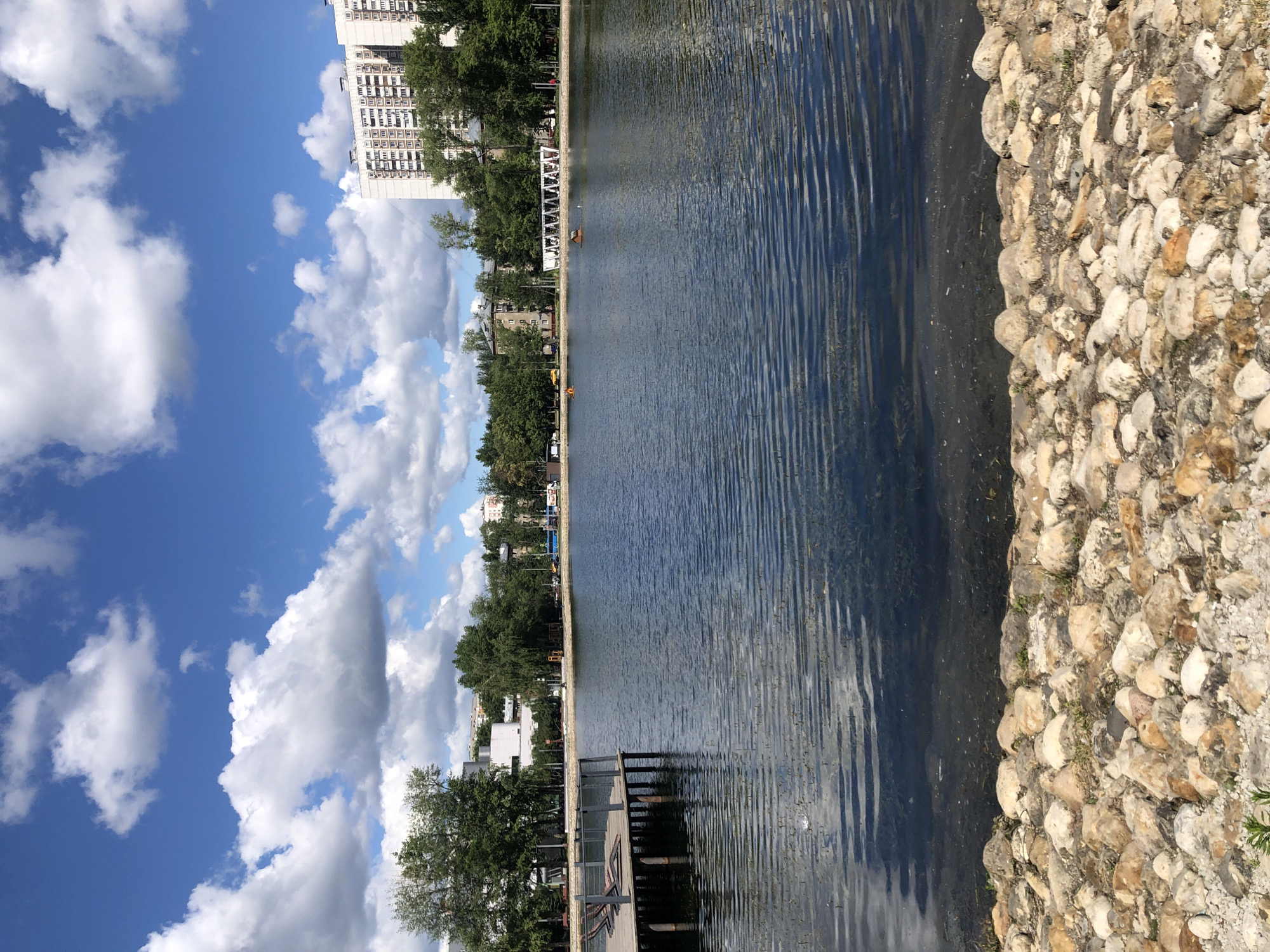 This is a photograph of a city park in Moscow, Russia. The image captures a serene waterscape with a lake or pond in the foreground, its surface rippled by a gentle breeze. The right side of the frame is bordered by a natural embankment made of large, irregularly shaped stones, with patches of green grass growing between them.
On the left side of the water, a wooden pier extends into the lake, featuring a railing and the suggestion of a walkway. Beyond the pier, a line of lush green trees forms a verdant backdrop. Further back, apartment buildings with a uniform architectural style are visible, suggesting an urban setting within the park. The sky above is a vibrant blue, dotted with fluffy white clouds, indicating a pleasant day, likely in the late morning or early afternoon. There are no people clearly visible in the frame, adding to the sense of peaceful solitude. The overall impression is one of a well-maintained urban green space, offering a tranquil escape from the city.