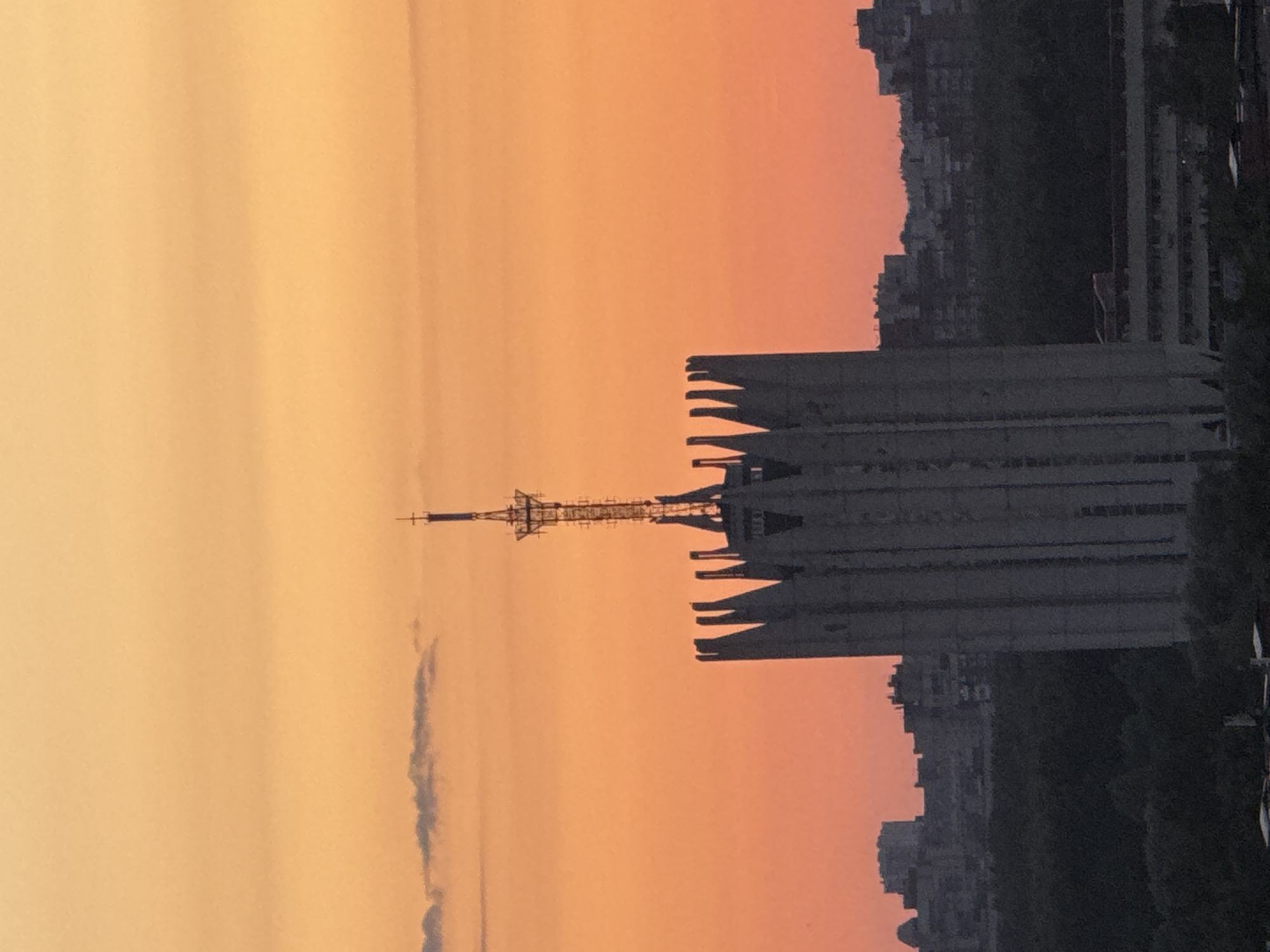 The image displays a panoramic urban landscape at either sunrise or sunset, characterized by a vibrant orange and yellow sky with a single elongated cloud in the upper left. Dominating the scene is a tall, brutalist-style building, dark grey in hue, featuring a distinctive, jagged, tooth-like top. From the center of this structure extends a slender, grey lattice communications tower equipped with multiple antenna arrays and topped with a small cross. The foreground features a dark, dense expanse of trees. In the background, a sprawling cityscape of numerous multi-story buildings, mostly dark and silhouetted, stretches across the horizon. The scene is located in the city of Unknown, Russia. No people or specific actions are visible.