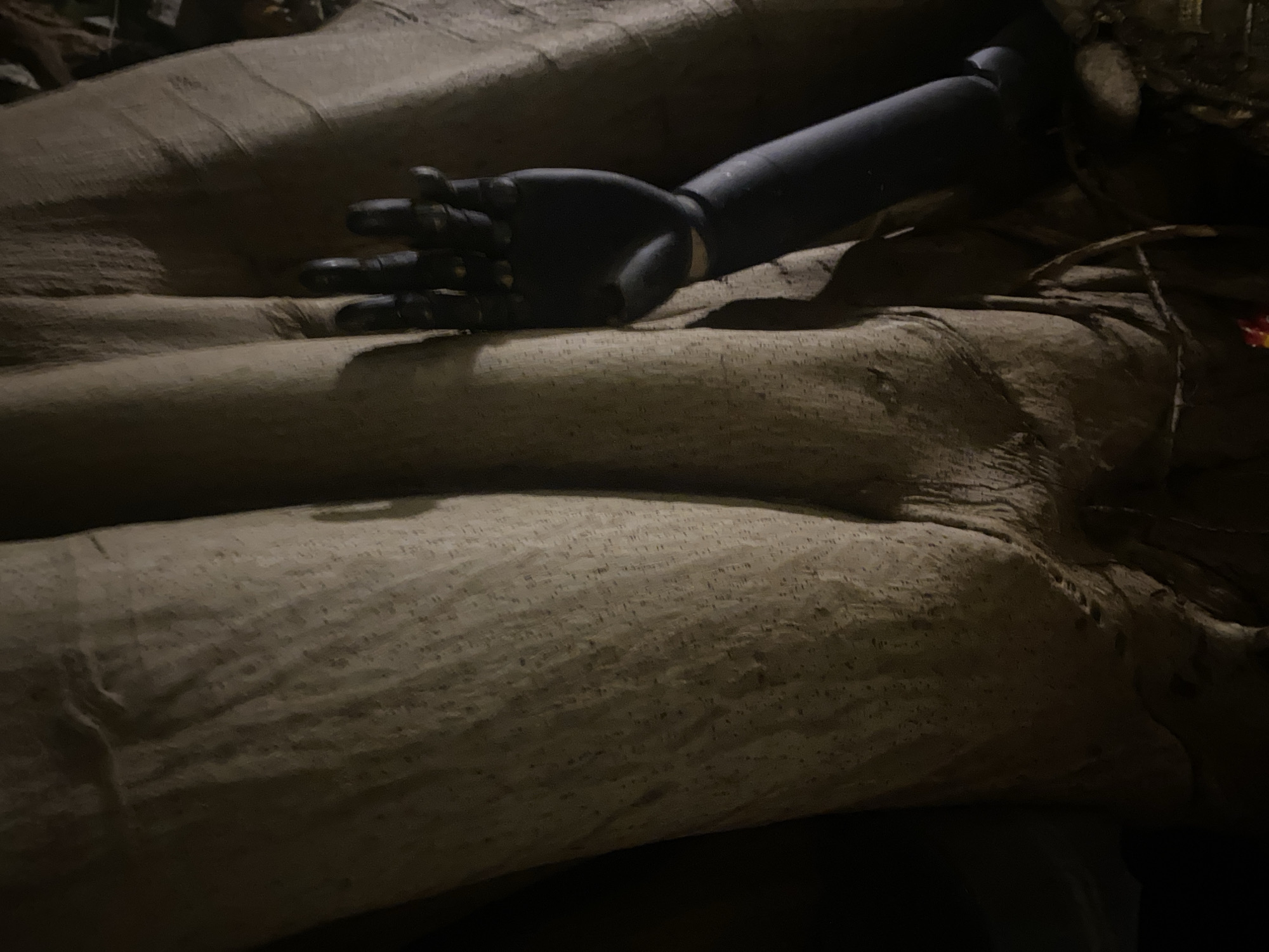 A close-up shot depicts a mannequin's arm and hand resting on what appears to be a burlap-covered surface. The arm is black and made of plastic. The burlap material has a coarse texture. The lighting is dim, casting shadows across the scene. The location is Baan Don Sai, Thailand.
