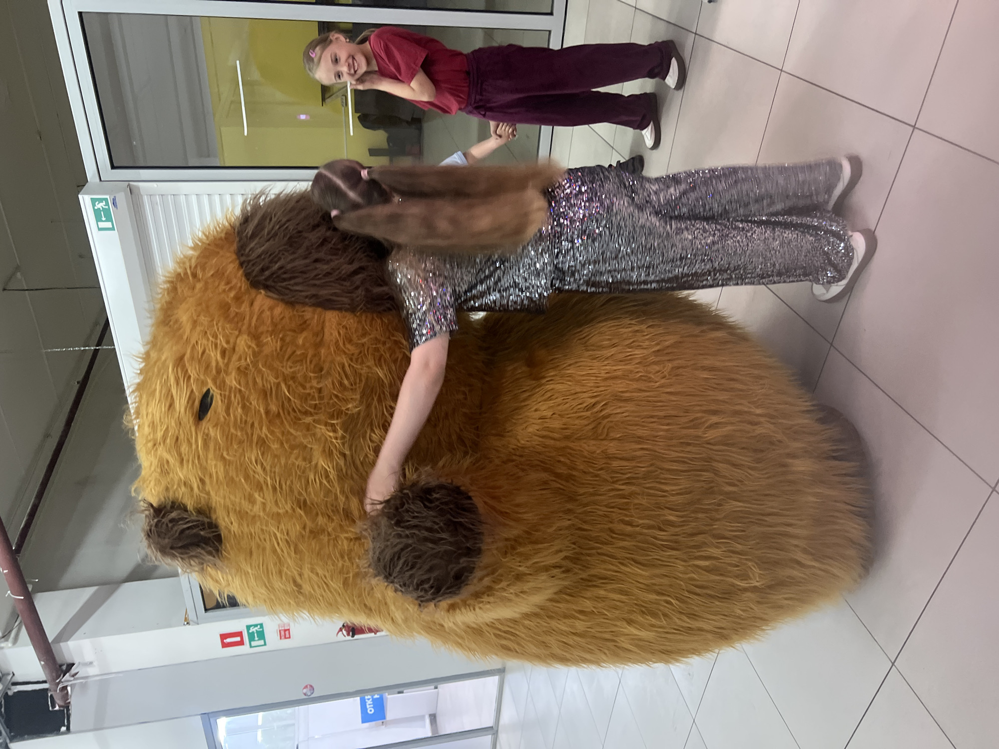 An indoor scene features two individuals interacting with a large, plush toy. In the foreground, a person with long, straight brown hair, possibly a girl or young woman, is embracing a substantial, roundish plush figure covered in shaggy, light brown fur. The plush toy has darker brown patches suggesting ears and a snout. The person hugging it wears a shimmering silver long-sleeved top and matching wide-leg trousers, paired with white-soled sneakers.
In the background, a younger girl with blonde hair tied in pigtails with pink hair ties stands behind a clear glass partition, smiling brightly towards the camera. She is dressed in a maroon t-shirt and matching pants, also wearing white-soled sneakers. Her hands are resting near her chin.
The setting appears to be a commercial or public indoor space, characterized by a white tiled floor with dark grout lines, and white walls or partitions. Behind the girl in the background, there's a large window or glass door with a white frame. Visible on the frame are several signs, including a green "exit" symbol (a running man) on a white background, and a red arrow pointing left. Further down, a blue sign has the Cyrillic text "ОТКР" visible, likely part of "ОТКРЫТО" (meaning "OPEN"). The overall atmosphere seems cheerful and casual.