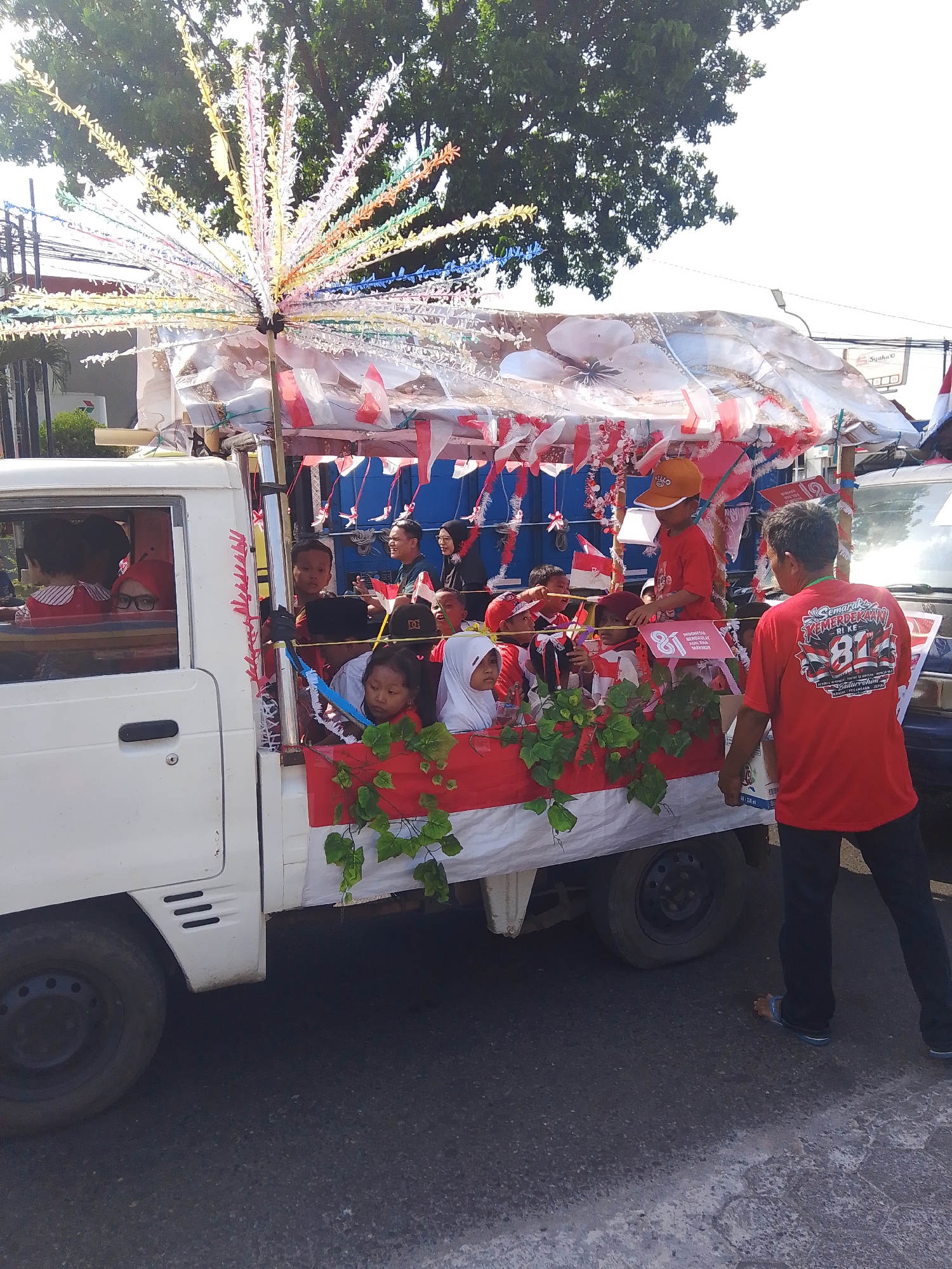 In Rejosari, Indonesia, a decorated flatbed truck is participating in a parade, likely for a national celebration. The truck is adorned with red and white flags, a common display of Indonesian patriotism. A cluster of colorful, star-shaped lights is mounted on a pole at the front of the truck's canopy. The truck bed is filled with children, many wearing red and white attire, and some wearing headscarves. A man in a red t-shirt with a graphic design on the back is standing at the rear of the truck, facing away from the camera, and appears to be overseeing the children. The truck is moving on a paved road under a clear sky, with trees visible in the background.