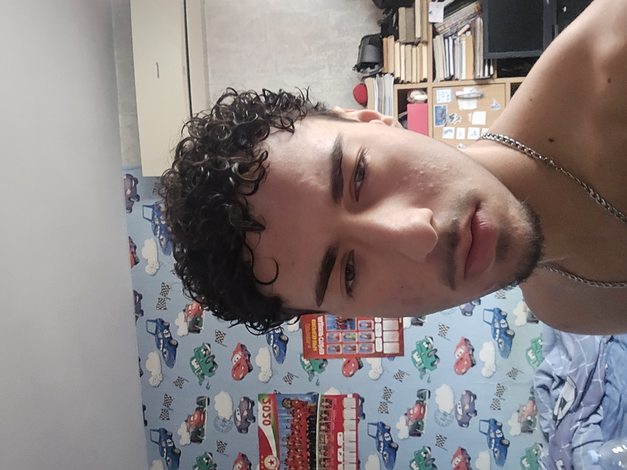 The image depicts a young adult male in what appears to be a bedroom located in Sofia, Bulgaria. The male subject occupies the center-foreground, facing generally forward with his gaze directed downwards and slightly to the viewer's left. He has dark, tightly curled hair, dark eyebrows, and sparse facial hair forming a goatee. He is shirtless, wearing a silver-colored link chain necklace. Minor blemishes are visible on his forehead and cheek.
The background reveals a domestic interior. The wall to the left of the subject is covered with light blue wallpaper patterned with cartoon racing cars (red, blue, green, and purple), white clouds, and black and white checkered flags. Two posters are affixed to this wall: one displaying a group photograph, a red banner with stars, the text "OSOS," and a graphic of a red double-decker bus; another, partially visible poster below it, shows a grid of small headshot photographs and includes partially legible Cyrillic text. A blue and white patterned duvet is visible at the bottom left, suggesting a bed.
The wall to the subject's right features a textured, light-colored surface. A beige-colored split-unit air conditioning system is mounted high on this wall. Below the air conditioner, a wooden shelving unit or bookcase is visible, containing multiple stacks of books with various spine colors, a black object (possibly a camera or device), and a red object (possibly a cap or small container). A black computer monitor is visible at the bottom right corner of the frame. A small corkboard or similar display with pinned items is also partially visible on the shelving unit.