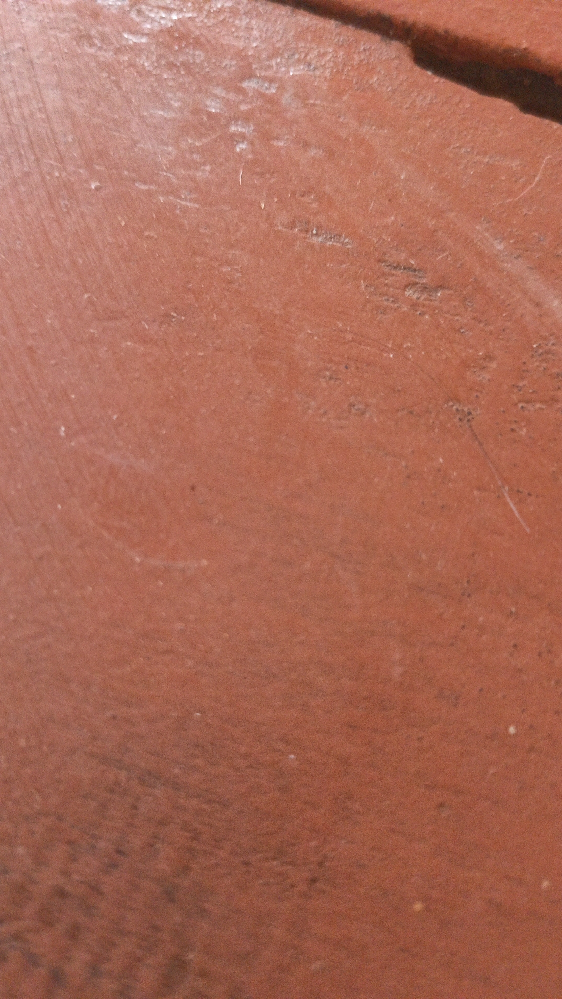 A close-up, overhead shot of a textured, reddish-brown surface with subtle variations in color and light. The surface appears to be somewhat worn, with faint scratches and scuffs visible. There are also small, scattered white specks and darker, granular areas. A dark, angular shadow dominates the top right portion of the image, suggesting an overhead object or structure. The overall impression is one of an aged, outdoor material, possibly concrete or stone, with a weathered patina. The image is a still shot, with no discernible action or people present. Given the provided location, this surface could be a portion of a road, sidewalk, building exterior, or other urban infrastructure in Ngembalrejo, Indonesia.