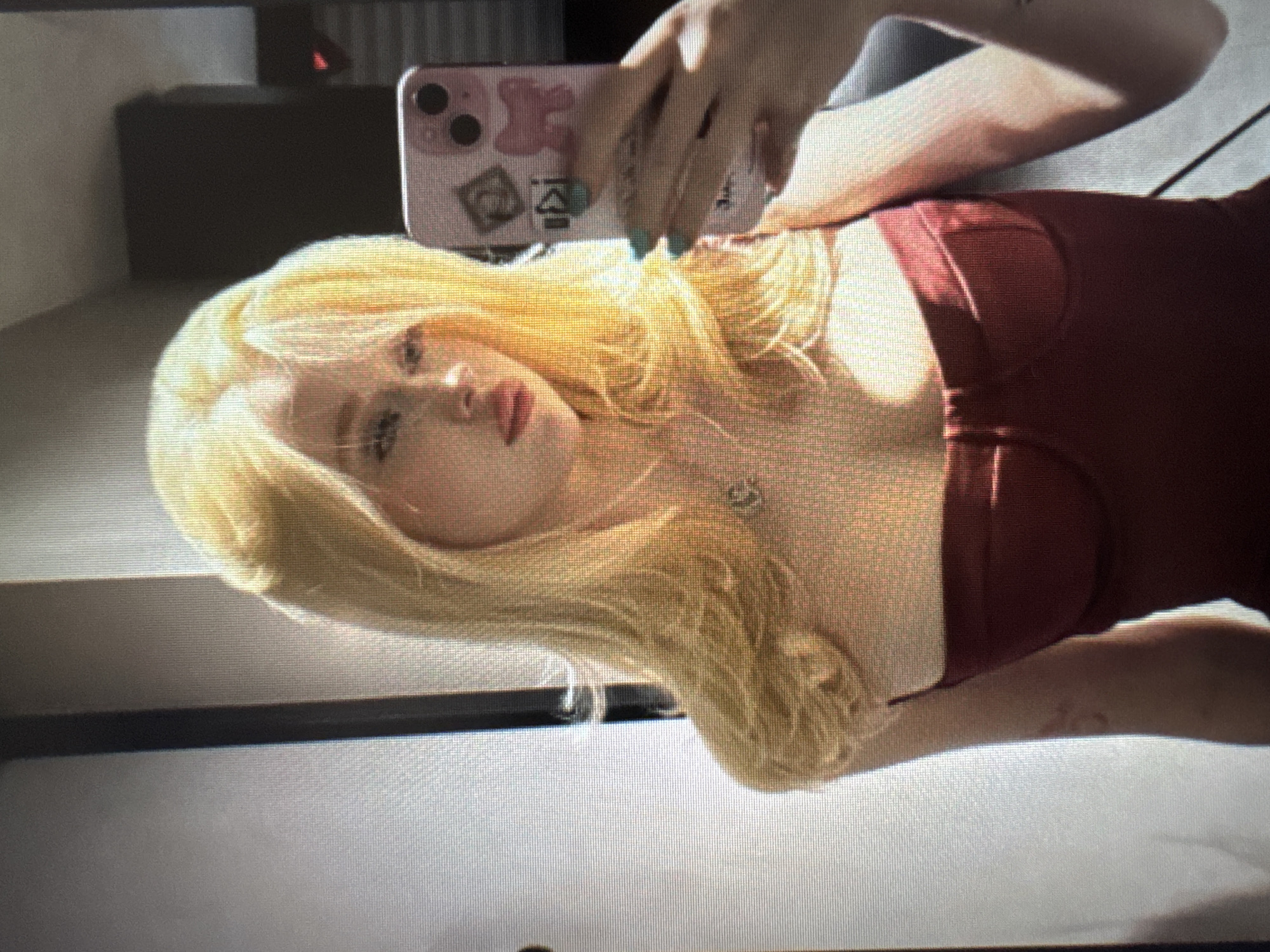 A blonde woman takes a selfie in an indoor setting, captured in an image that appears to be a photograph of a screen, showing distinct pixelation. She is positioned horizontally in the frame, with her head towards the left and her body extending to the right. Her long, bright blonde hair frames her face, and she has fair skin, with visible red lipstick.
She holds a light pink or lavender smartphone with her right hand, which has nails painted in a light teal or mint green. The phone case is adorned with several stickers, including a pink teddy bear and a green diamond-like shape, and another less discernible black and white symbol. The phone’s dual camera lenses are visible. Around her neck, she wears a silver necklace with a pendant. She is dressed in a reddish-brown or maroon strapless top or dress.
The woman's gaze is directed towards the phone's camera, with a neutral to slightly posed expression. Natural light illuminates her from the right, casting shadows and highlighting her features. The background is softly blurred, showing a dark area behind her and a lighter surface in the foreground, possibly a wall, furniture, or bed. The image captures a casual, self-portrait moment. No explicit text is clearly readable on the phone case stickers.