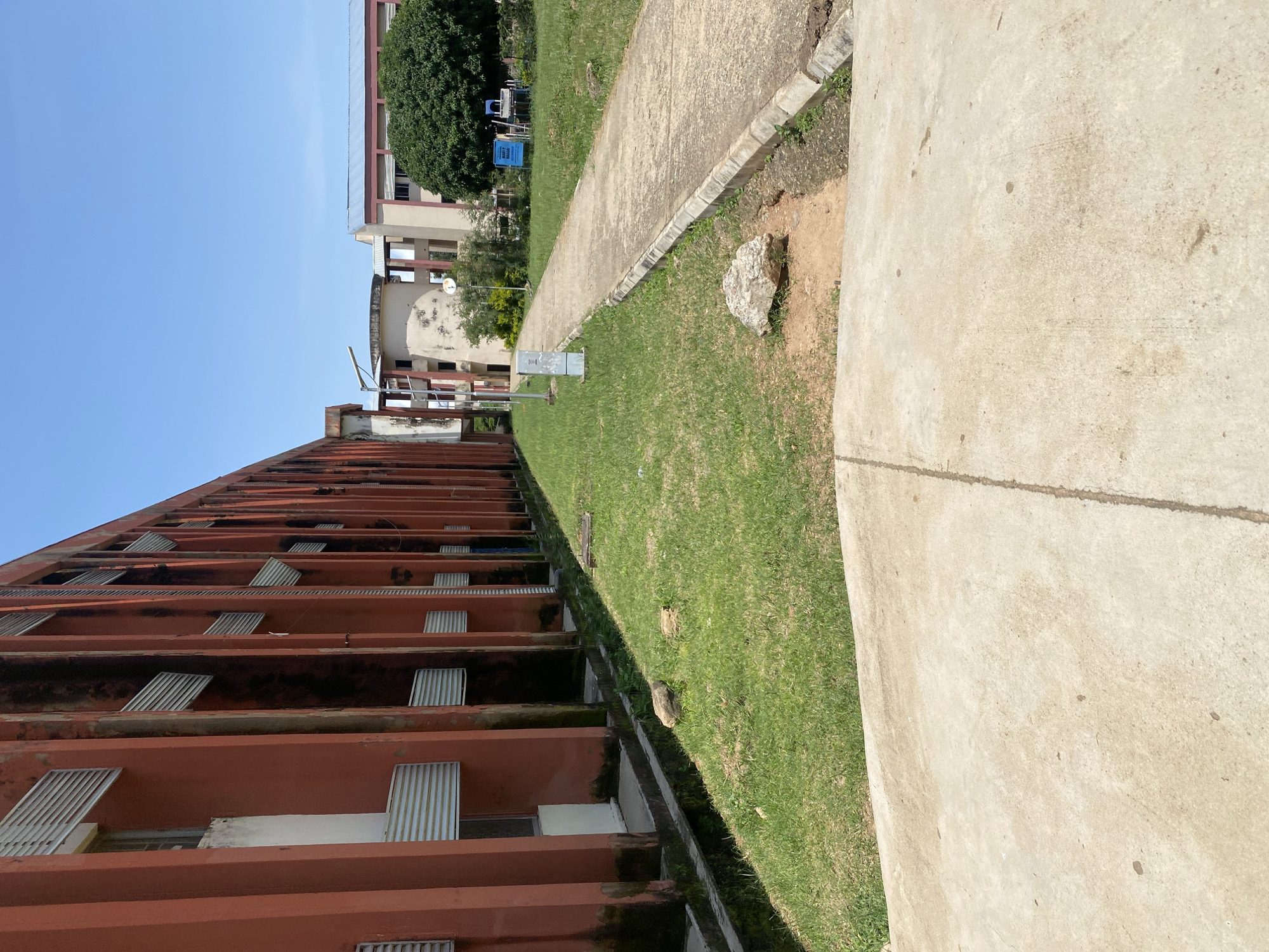 A low-angle, full-shot photograph captures a view of a long, reddish-orange building with vertical columns and horizontal slatted windows. To the right of the building, a concrete pathway winds through a grassy area. In the background, other buildings with lighter facades and a large, green tree are visible under a clear blue sky. The scene appears to be an outdoor area on a university campus in Jos, Nigeria, possibly the University of Jos, given the architectural style and the presence of academic-looking buildings. The pathway is bordered by a concrete curb, and the grassy area features a few scattered rocks and a small electrical box. The overall impression is one of a bright, sunny day on a campus with established, somewhat weathered architecture.