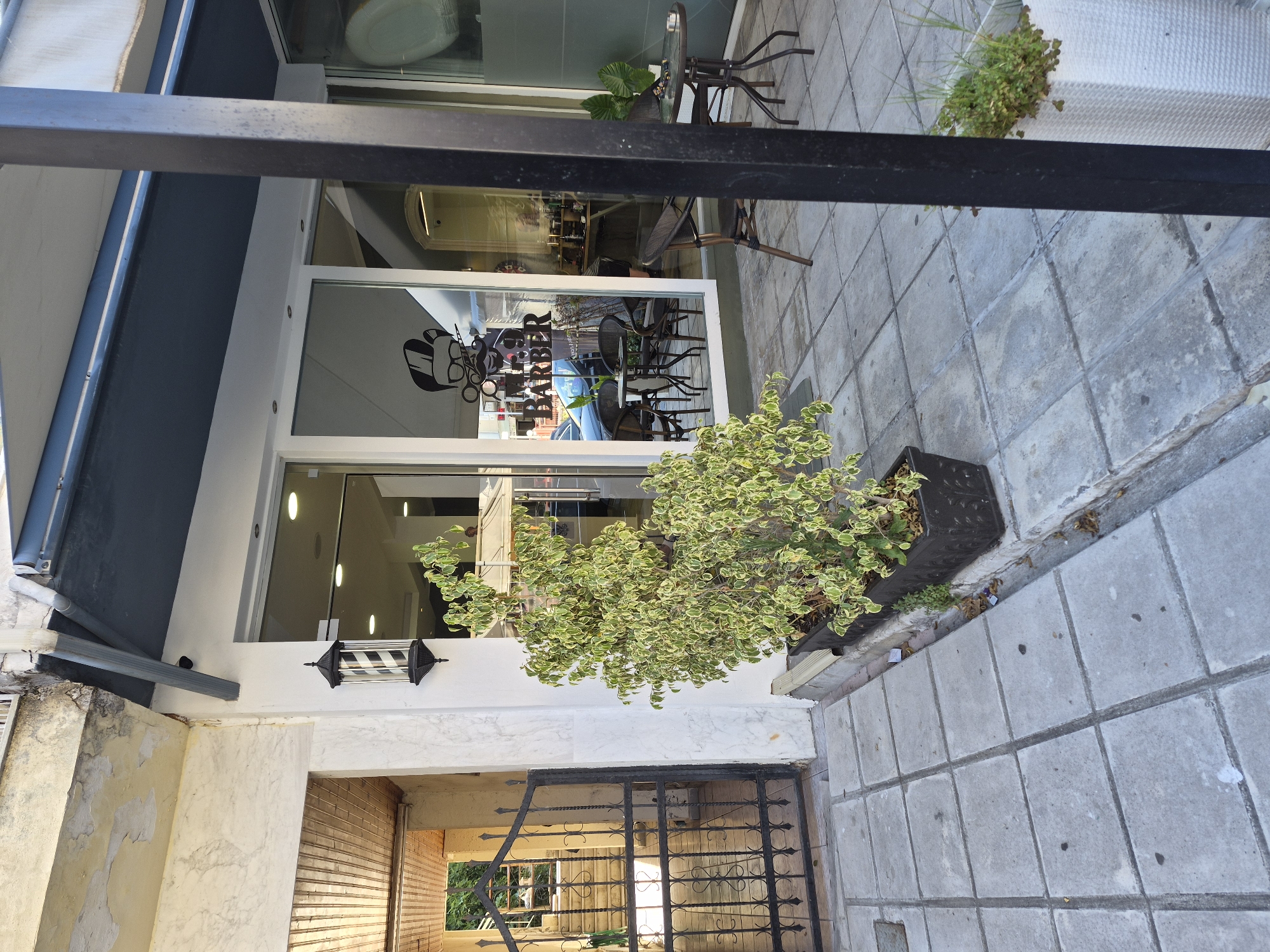 The image depicts the exterior of a barber shop in Nea Kallikratia, Greece. The shop features a large glass storefront with a black awning above. A stylized graphic of a barber with a mustache and scissors is visible on the glass, along with the text "MR BARBER". Inside, tables and chairs are arranged, suggesting an outdoor seating area.
In front of the shop, a decorative barber pole with black and white stripes is mounted on the wall. A potted plant with variegated green and white leaves sits in a black planter on the tiled sidewalk. To the left of the barber pole, a black metal gate provides access to a stairway leading upwards, with a wooden ceiling visible.
The sidewalk is paved with square concrete tiles. A black metal pillar stands to the right of the frame, partially obscuring the view of a small round table with two chairs and another potted plant in a white woven planter. The overall scene suggests a commercial establishment on a street with a combination of old and modern architectural elements.