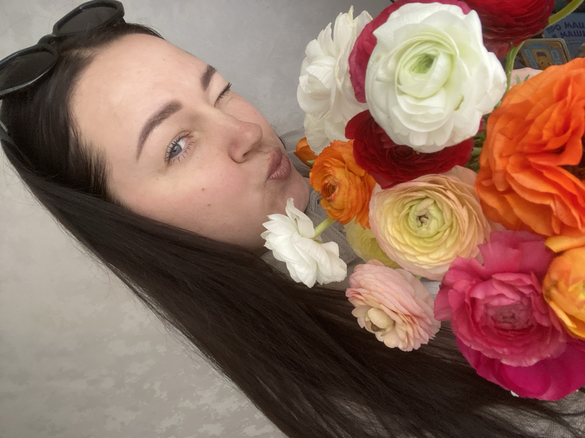 A woman with long, dark hair is positioned at an angle facing left, with her head slightly tilted. She is puckering her lips and has sunglasses perched atop her head. She is holding a bouquet of colorful ranunculus flowers that feature shades of white, red, orange, yellow, and pink. A book in Russian text can be seen in the background, partially obscured by the flowers. The environment appears to be indoors with a wall in the background with a blurred pattern of light and shadow. There is no indication of the location being Sochi, Russia.