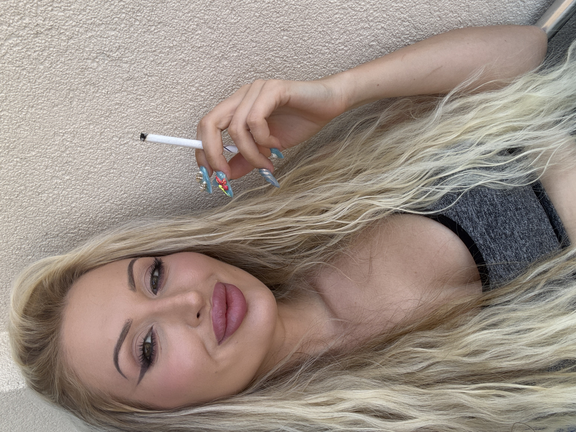 A woman with long blonde hair and heavy makeup is holding a cigarette between her fingers, with her nails painted in a colorful, elaborate design. She is wearing a grey sports bra and is leaning against a textured, light-colored wall. The setting appears to be outdoors, possibly on a balcony or patio.