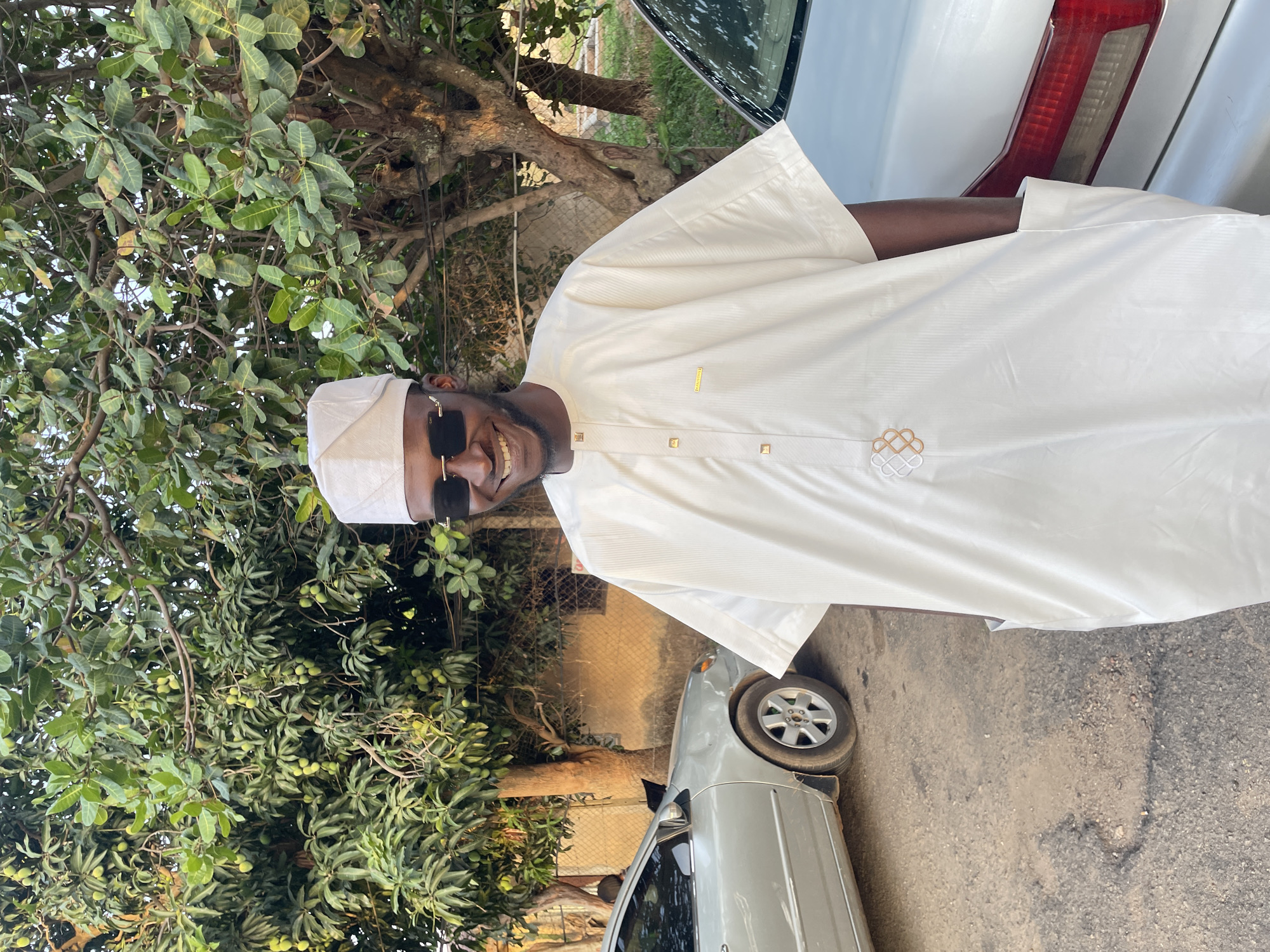 A man wearing a white traditional outfit, a white hat, and dark sunglasses stands next to a light-colored car on a paved road. The car is positioned so that part of its rear is visible. Behind the man and the car is a leafy green tree, possibly a mango tree, with many small green fruits. A chain-link fence and a building can be seen in the background. The setting appears to be outdoors on a sunny day. Jos, Nigeria is the location.