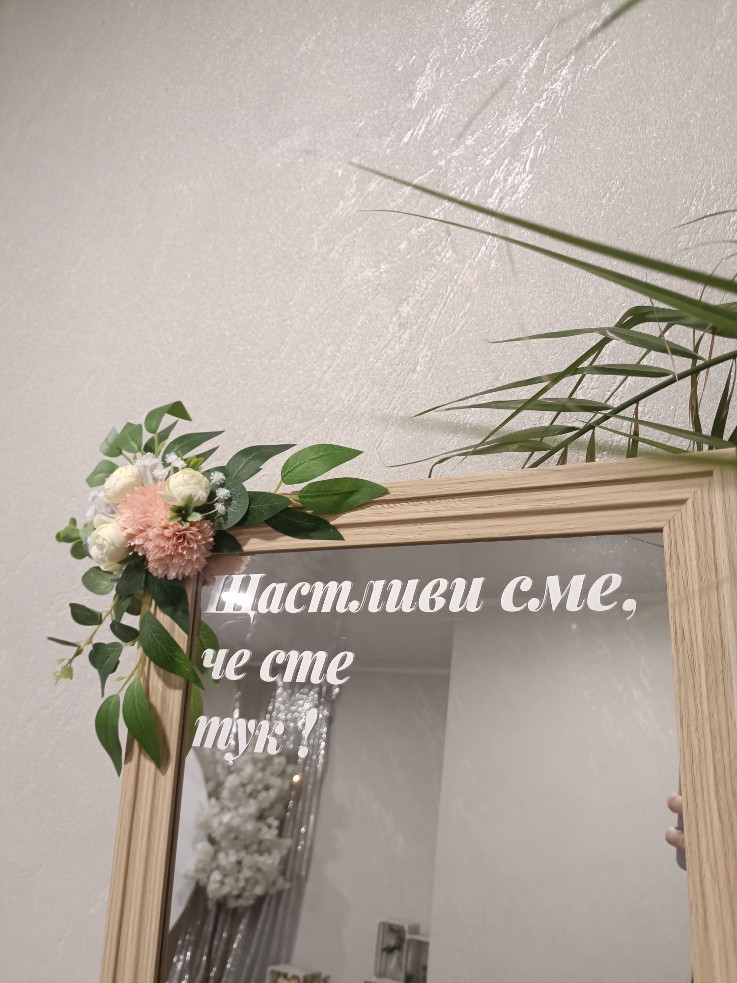 A framed mirror hangs against a textured white wall. The top left corner of the mirror is decorated with a floral arrangement of light pink, white, and green flowers and leaves. White text is written on the mirror glass. In the reflection of the mirror, another floral arrangement of white flowers and a silver curtain can be seen. A plant is located next to the frame on the right. A hand can be seen near the right side of the mirror. The image was taken in Plovdiv, Bulgaria.