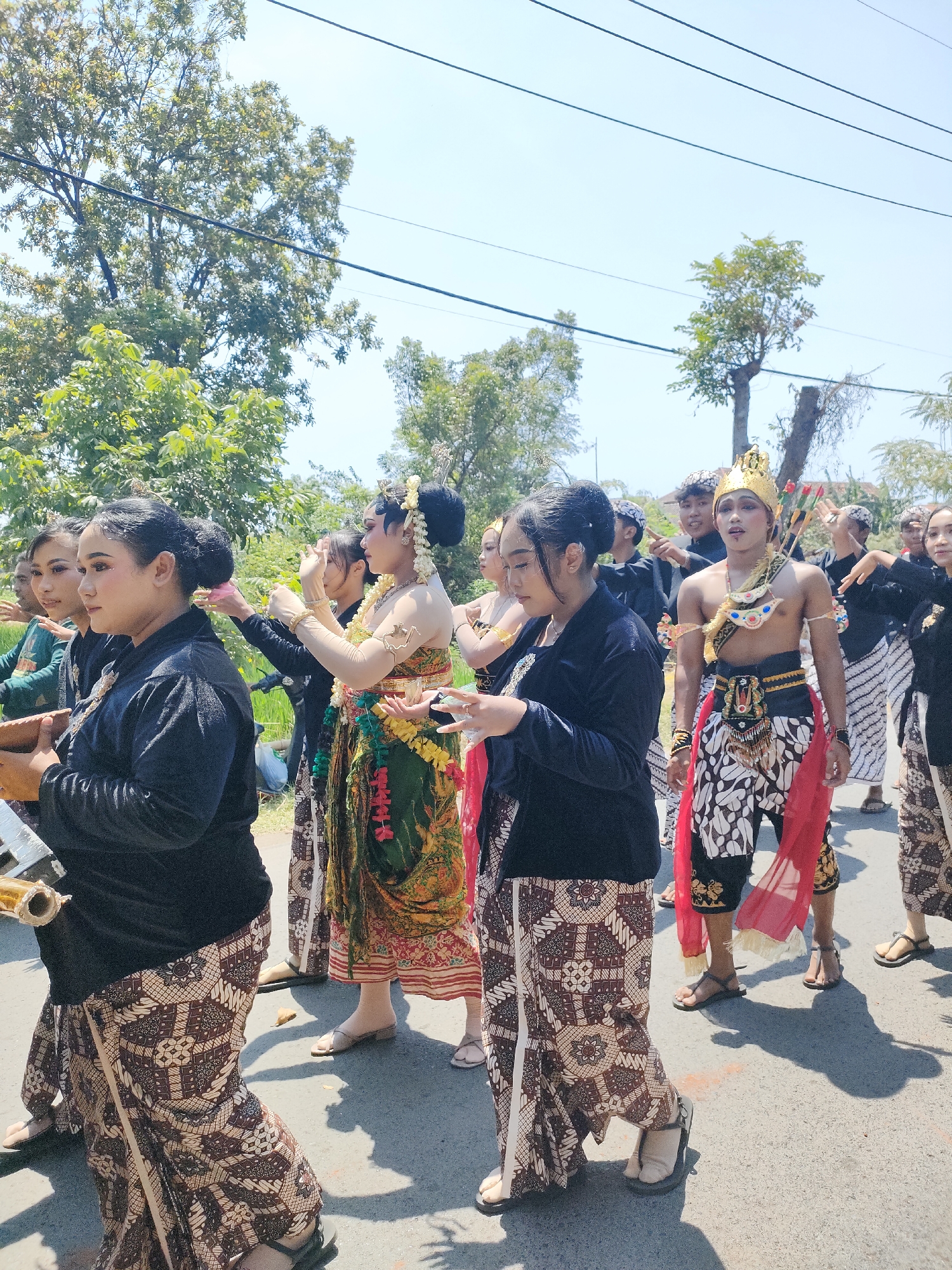 This is a medium shot, eye-level, vertical photograph captured in Tongaswetan, Indonesia. The image depicts a procession of people dressed in traditional Indonesian attire walking along a paved road under a clear, bright sky. The participants appear to be part of a cultural performance or ceremony.
In the foreground, on the left, a young woman in a black long-sleeved top and a patterned brown batik skirt is holding a cylindrical object. Her hair is styled in an updo, and she wears makeup. Beside her, to her right, another woman with similar attire and hairstyle is partially visible. Further to the right, a woman in a green and gold ornate traditional costume with a floral pattern and a garland of flowers around her neck is visible. She is also adorned with elaborate jewelry. Her hair is styled in a bun with decorative hairpieces. She is gesturing with her hands, palms open, as if performing.
The central figure is a young man dressed as a warrior or king. He wears a golden crown with ornate embellishments, a chest piece, and a patterned loincloth with a red sash draped over his shoulders. He has accessories on his back, possibly arrows or spears, and is adorned with jewelry. He is looking forward with his mouth slightly open.
Other participants, mostly women in black tops and batik skirts, and a few young men in black tops and patterned sarongs, are seen walking behind. They are all facing forward, participating in the procession. Some of them are holding objects or making hand gestures.
The environment includes lush green trees and foliage on the left side of the road, with power lines running overhead. The road is asphalt, and shadows of the participants are cast on the ground. In the background, more trees and some buildings can be vaguely discerned. The atmosphere appears to be festive and celebratory, with the bright sunlight illuminating the scene.