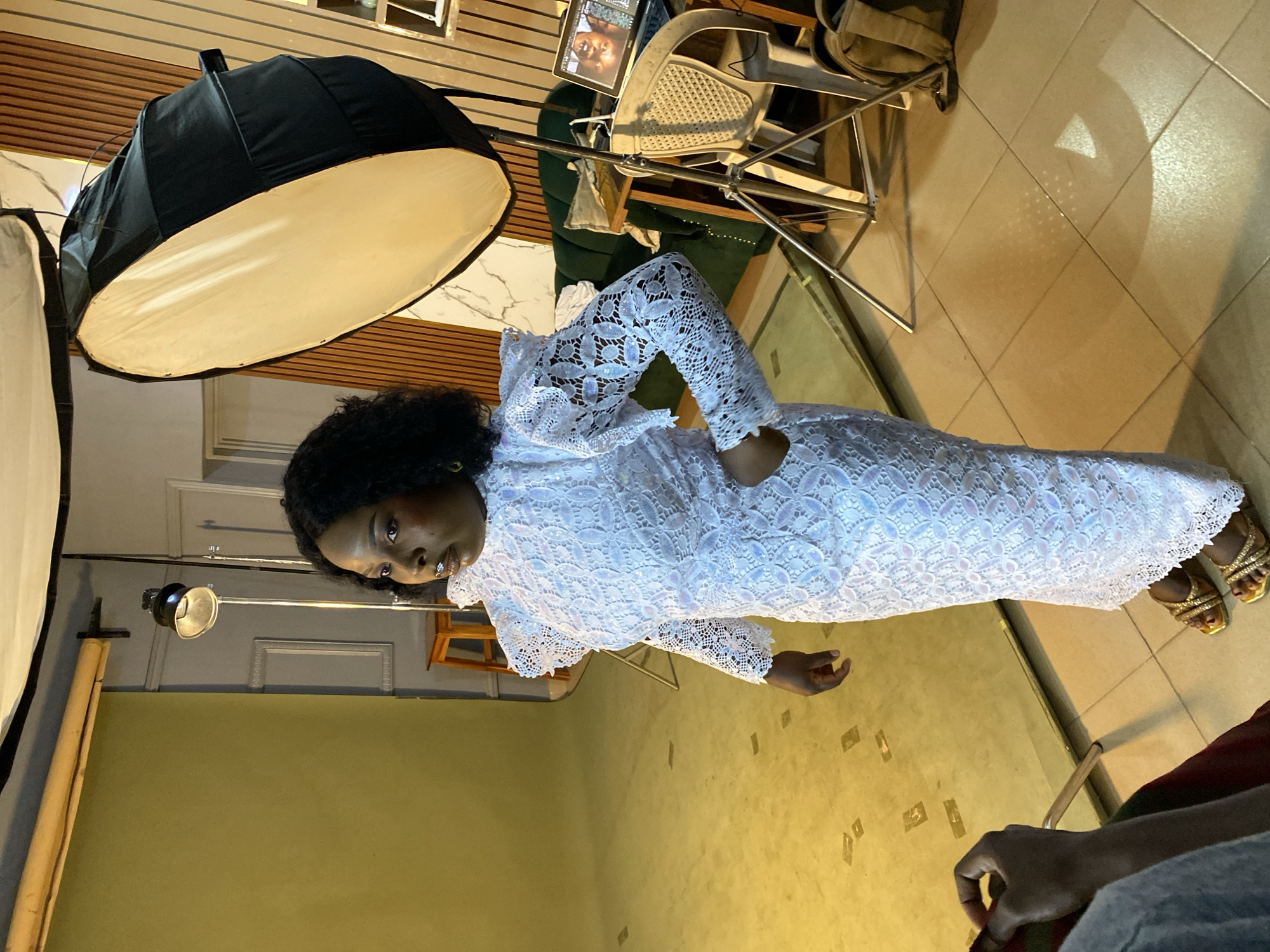 The image captures an indoor scene, likely a photography studio or a dedicated space for a photoshoot, given the presence of professional lighting equipment. The primary subject is a woman with dark, curly hair, standing and looking towards the camera with a calm, posed expression. She is wearing an elegant, long white dress with intricate lace or sequin patterns, and shiny gold high-heeled sandals.
In the background and surrounding her, various objects associated with photography are visible. To her left, a large black softbox with a white diffuser is prominent, mounted on a stand. Another smaller light fixture is also present. Further back, a light-colored chair, a metallic stand, and a dark bag are seen. A laptop is placed on a surface behind her, displaying an image of a person's face on its screen. The floor is made of light-colored tiles, and shadows from the equipment are cast upon it. The walls feature different textures; one section appears to be a plain olive-green color, while another has horizontal wooden paneling with white, marble-like accents.
The activity taking place is clearly a photoshoot, with the woman as the model. Another person's hand is visible in the bottom right corner of the frame, possibly operating a camera or assisting with the shoot. The overall lighting suggests artificial illumination typical of a studio setting. No visible text can be deciphered in the image. The scene conveys a professional and staged environment for creating photographic portraits.
