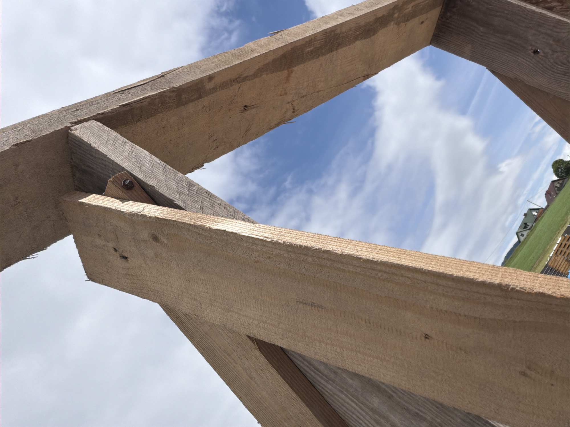 The image is a low-angle, close-up shot of wooden structural beams against a cloudy sky. The wood appears to be unfinished lumber, with visible grain and rough edges. One beam has a bolt and nut securing it to another piece of wood. In the background, a green, rolling landscape is visible with some houses and trees. The scene is likely outdoors, possibly a construction site or a rural setting in Męcina, Poland. There are no people or animals in the image. The focus is on the texture and construction of the wooden beams.