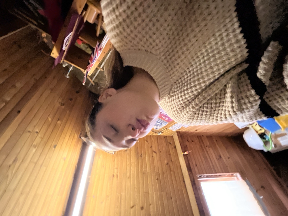 The image depicts the interior of a room in a dwelling located in Ancenis, France. The scene is rotated approximately 45 degrees clockwise, presenting the subject and room features at an oblique angle.
The primary subject is a female, appearing to be a young adult, with fair skin and dark hair, visible from the chest upwards. She is wearing a light beige or cream-colored waffle-knit sweater featuring two dark horizontal stripes across the lower arm and torso area. Her eyes are closed, and her lips are pursed into a pronounced pout.
The room is characterized by extensive light-colored wooden paneling covering the walls and ceiling. A horizontal fluorescent light fixture is affixed to what would be the ceiling in a normal orientation, illuminating the upper portion of the frame. In the lower left quadrant of the rotated image, a window with a white frame is visible, admitting diffused natural light. Below the window, on what appears to be a surface or shelf, a white hard hat rests alongside other small, multi-colored objects, including blue and yellow items.
In the upper right quadrant of the rotated image, a wooden shelving unit is partially visible. The shelves hold various items, including textiles, what appears to be a clear glass bottle, and other unidentifiable objects. The overall lighting in this section of the room is dimmer than near the window and fluorescent fixture.