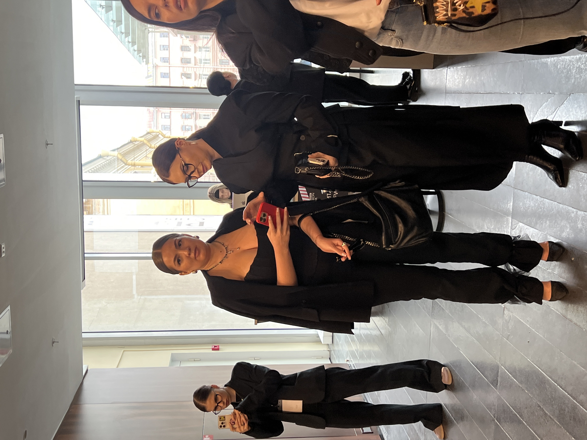 The image depicts a group of people indoors, likely attending an event or gathering. One woman is wearing an all-black outfit with a jacket draped over her shoulders, holding a red phone. Another woman wears glasses and a dark jacket, holding a dark-colored purse with a chain. A third person, also wearing glasses, is dressed in a black suit and using a white smartphone. Other individuals are partially visible, including one wearing a leopard-print purse. The location appears to be a modern interior space with large windows offering a view of buildings in what is possibly the city of Saint Petersburg, Russia.
