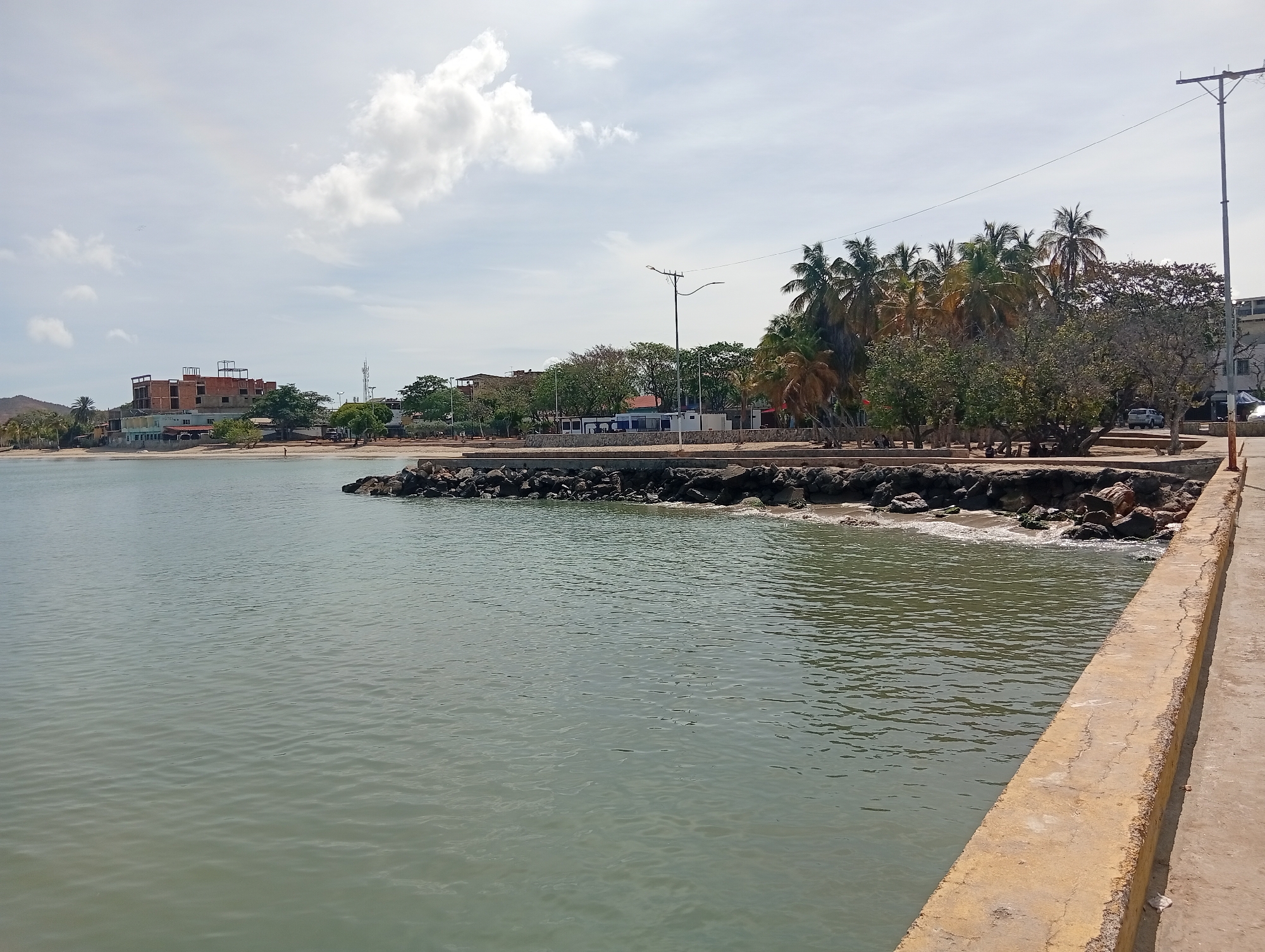 The image shows a view from a pier into a bay in Juan Griego, Venezuela. The water is calm and a greenish-gray color. A concrete pier stretches along the right side of the image. The shoreline is lined with a breakwater of rocks and boulders. Behind the rocks is a narrow beach area with trees, including palm trees. Buildings are visible along the shoreline, including a partially constructed building. A few people are visible in the distance on the beach. Overhead power lines are also visible. The sky is partly cloudy.