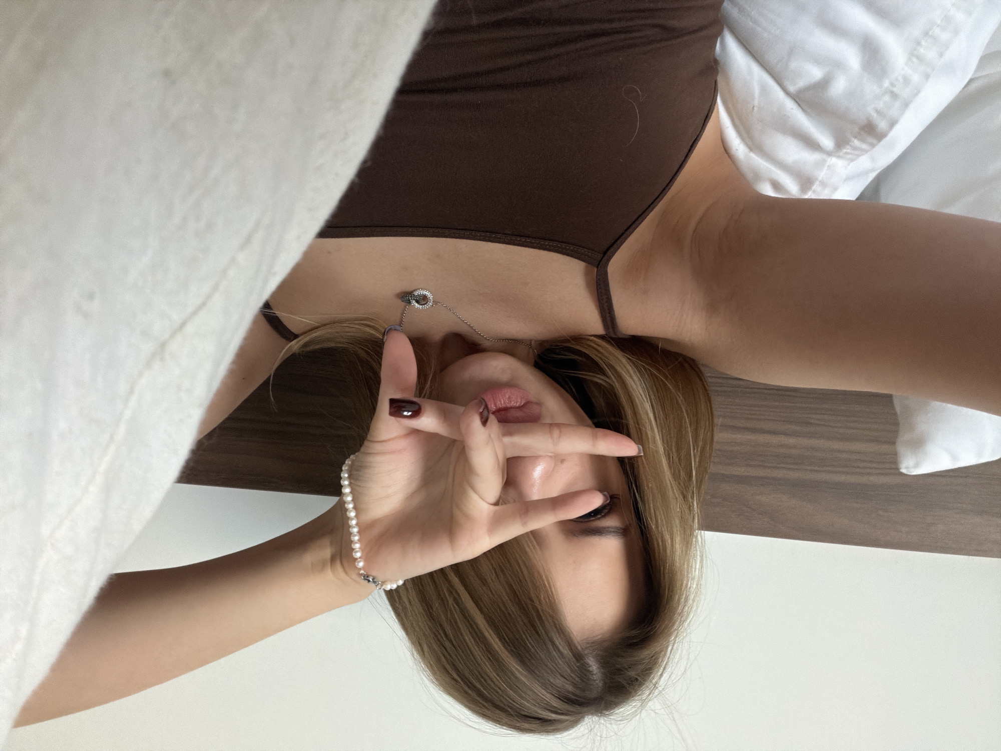 A young woman with blonde hair is lying on a bed, holding her hand up to her face in a gesture. She is wearing a brown tank top and a pearl bracelet. A silver necklace with two interlocking rings is visible around her neck. The bed has a wooden headboard and white pillows and bedding. The background is a plain white wall. There is no indication of the city of Đà Nẵng, Vietnam in this image.