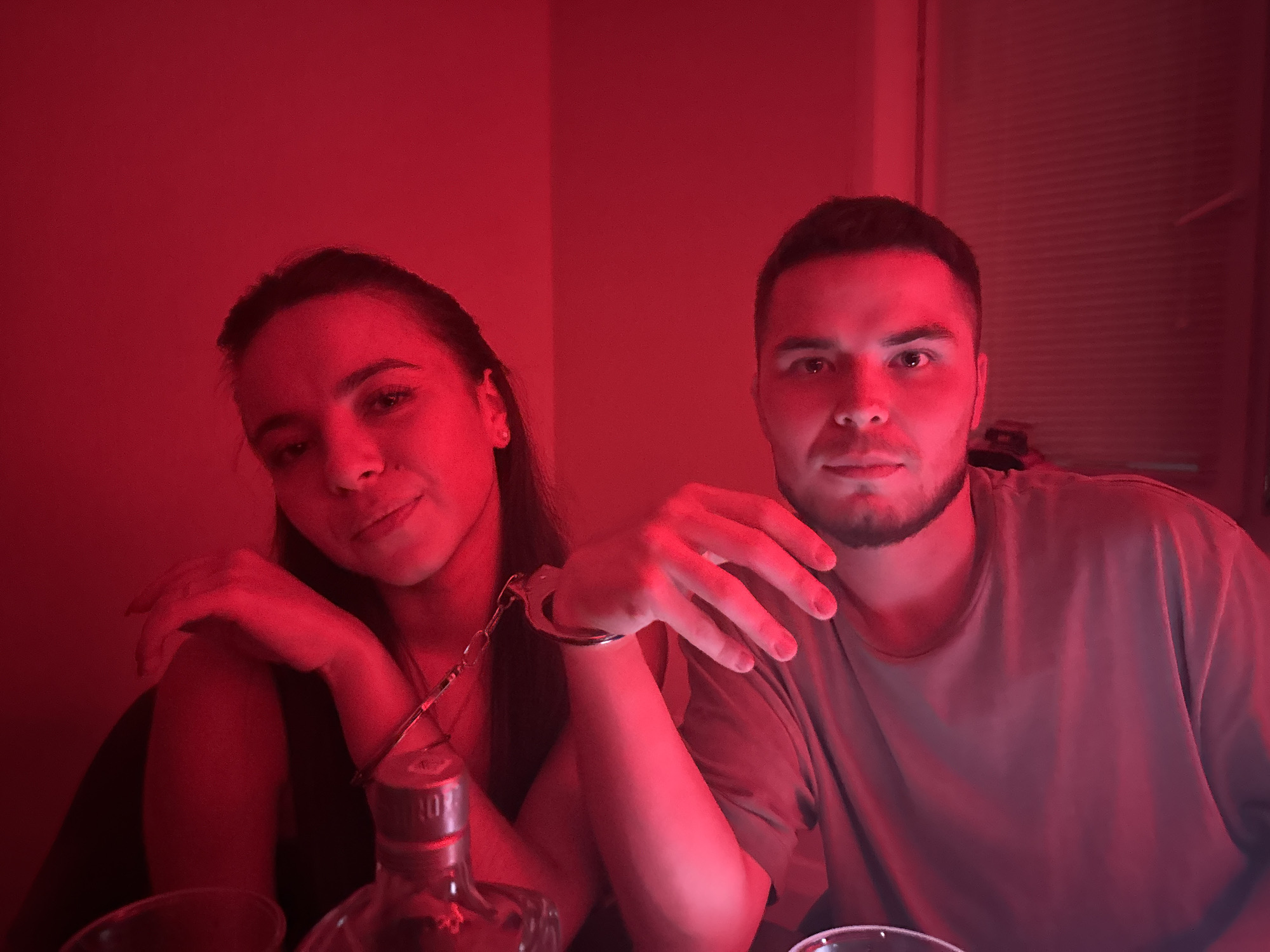 In a dimly lit room bathed in red light, a young man and woman are seated side-by-side, their wrists bound together by a set of handcuffs. The woman, on the left, has dark hair and is looking towards the camera with a slight smile. The man, on the right, has short dark hair and a beard, and is also looking at the camera. A bottle of alcohol with a silver cap is visible in the foreground. The background appears to be a wall with blinds. The overall impression is one of a staged or playful scene, possibly in a private residence or a themed establishment. The location of Perm, Russia, is not discernible from the image itself.