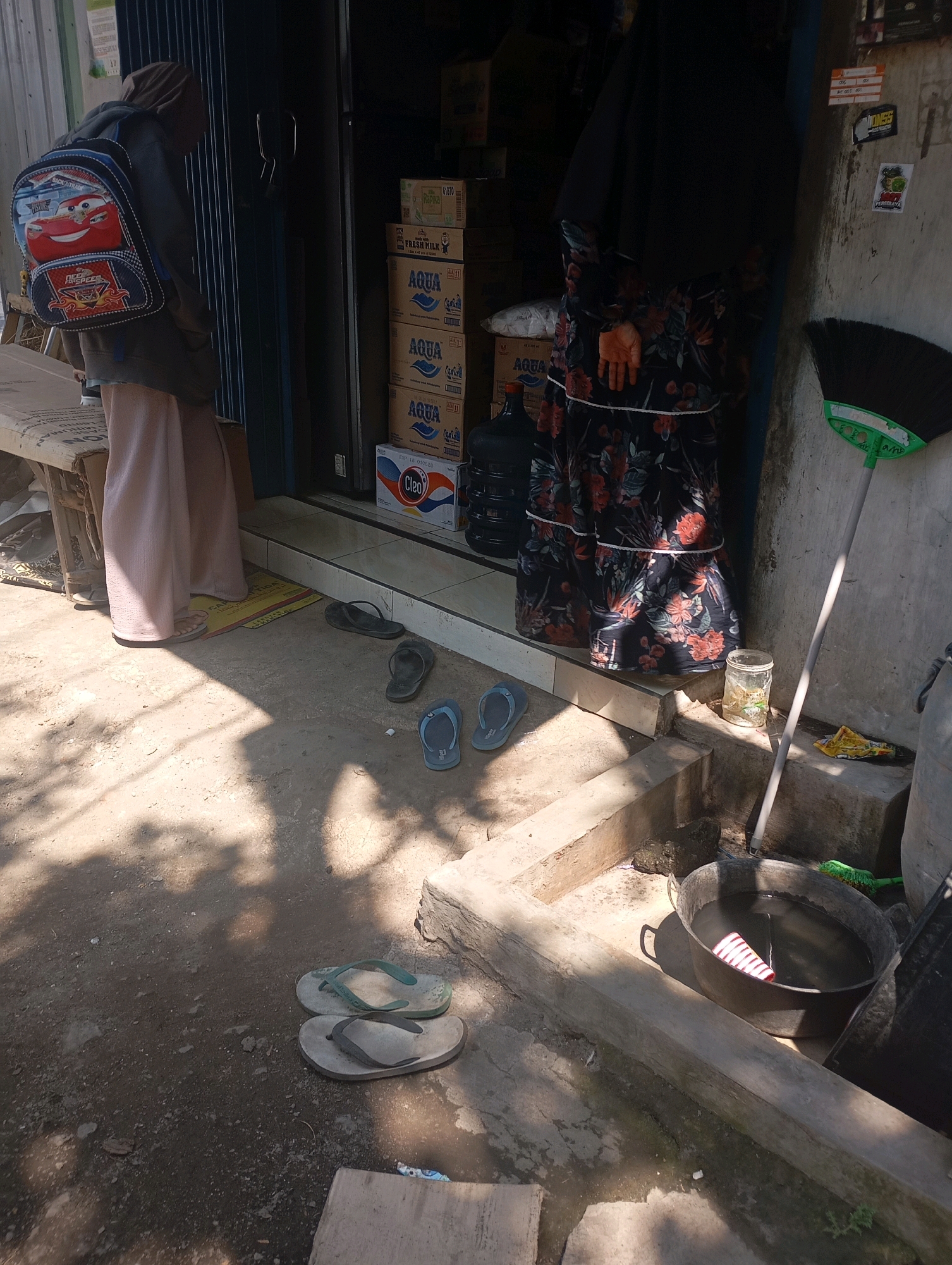 In Ketegan, Indonesia, a person wearing a hijab, a brown jacket, and light pink pants stands with their back to the camera, carrying a colorful backpack featuring the character Lightning McQueen. The person is facing a storefront with a dark metal accordion door. Stacked boxes of bottled water, labeled "AQUA," and a "Cleo" brand box are visible behind the door. Scattered on the ground in front of the store are several pairs of flip-flops and sandals. To the right of the entrance, a broom leans against the wall, and a large metal basin filled with dark water sits on a raised concrete platform. A small glass jar with some items inside is placed on the platform near the basin. The ground is a mix of dirt and concrete, with shadows cast by overhead foliage.