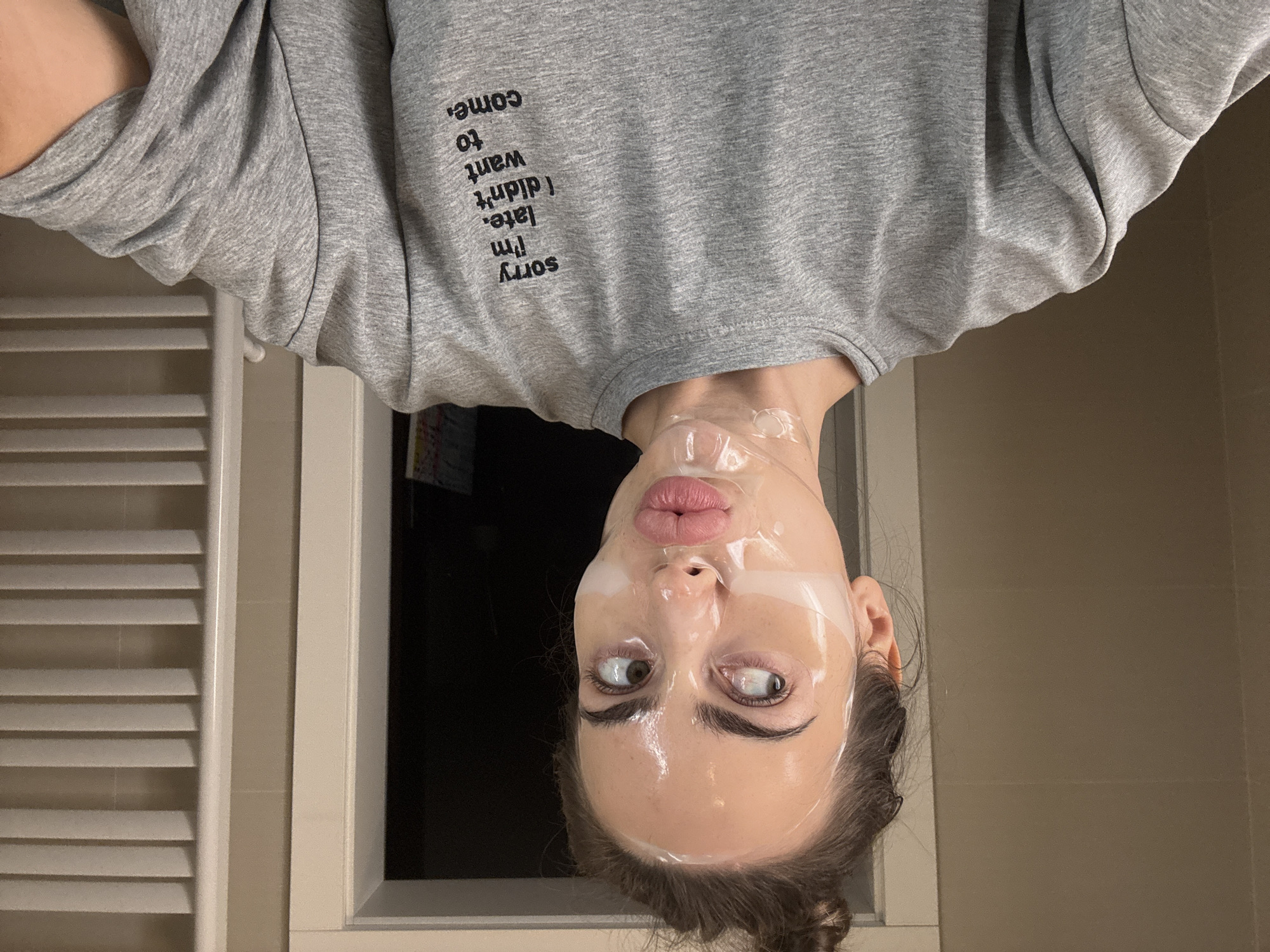 A photograph shows a fair-skinned individual with dark eyebrows and brown eyes, wearing a clear face mask. The mask covers their entire face. They are making a puckered expression. The individual is wearing a grey sweatshirt with text that reads "Sorry, I'm late, I didn't want to come" printed on the upper chest. The image appears to be rotated 180 degrees. In the background, there is a white radiator to the left and a dark window centered behind the individual's face. A light-colored tiled wall and trim are also visible. Without additional information, the city or country cannot be determined from the image.