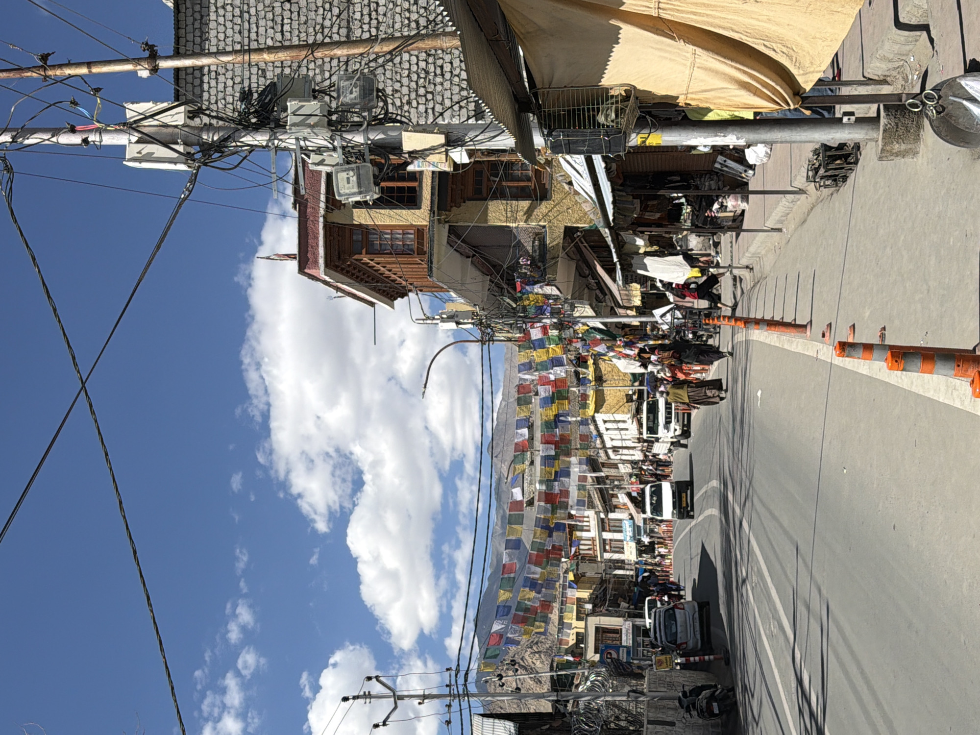 The image depicts a street scene in Leh, India, likely on the main market road, as indicated by the presence of shops, vehicles, and pedestrians. The sky is blue with scattered white clouds. Strings of colorful Tibetan prayer flags are strung across the street, adding a vibrant element to the urban landscape.
The street is paved and appears to be a busy thoroughfare with several vehicles, including cars, vans, and a motorcycle, driving or parked along its sides. Pedestrians are visible walking on the sidewalks and crossing the street. The buildings lining the street vary in architectural style, with some featuring traditional wooden balconies and multi-story structures. Shops with awnings are set up along the sides of the road, displaying various goods.
Utility poles with a complex network of wires and electrical boxes are prominent features, indicating the presence of infrastructure. The overall environment appears to be a typical bustling market area in a Himalayan town. The lighting suggests it is daytime, with the sun casting shadows on the road and buildings.