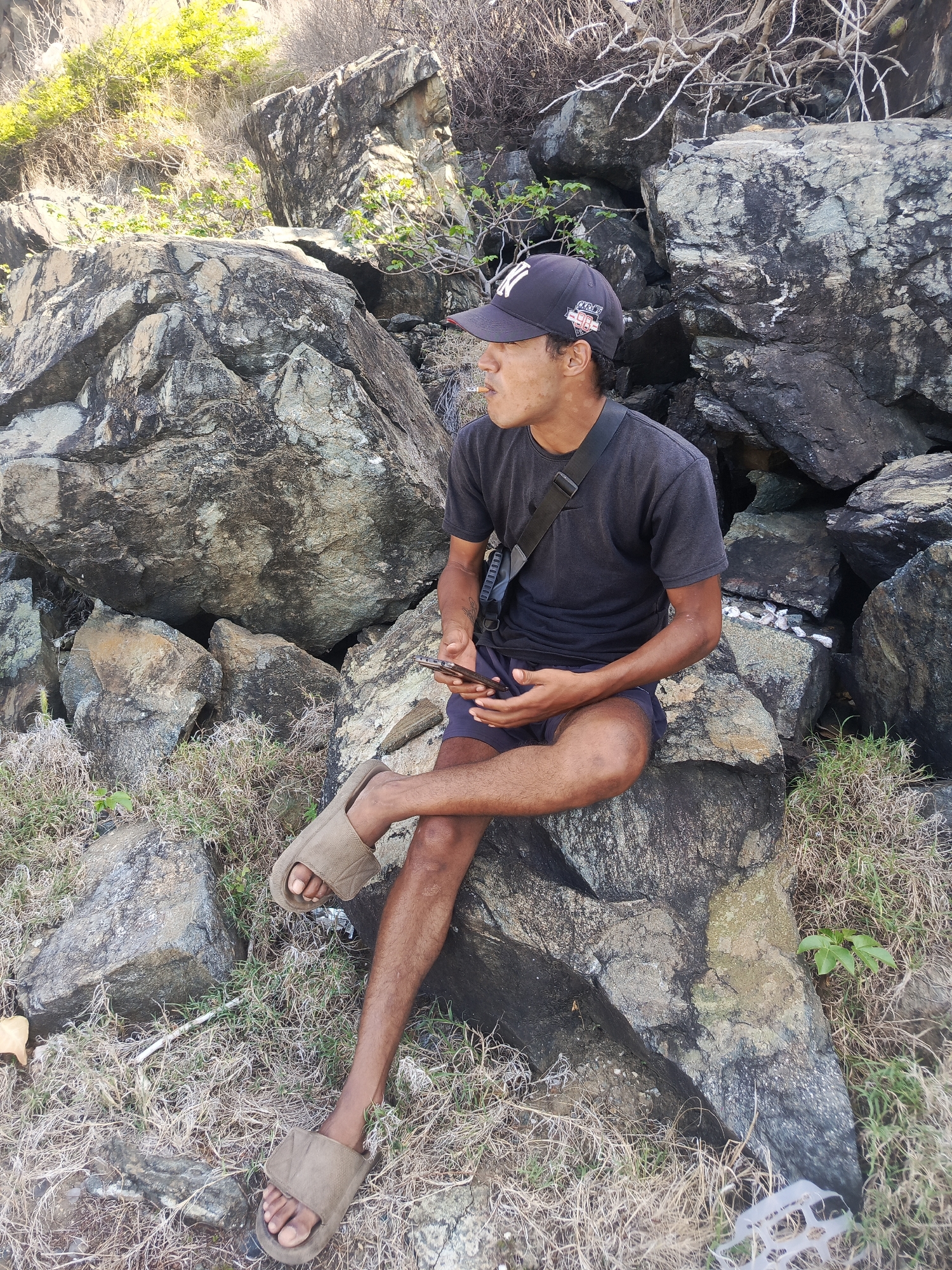 A young man sits on a large rock outcropping, holding a smartphone in his right hand and looking to his left. He is wearing a dark t-shirt, dark shorts, a dark baseball cap, and light brown slide sandals. A black crossbody bag is slung over his left shoulder. He appears to be smoking. The man is seated amongst a rocky, dry landscape with sparse vegetation, typical of the coastal areas of Pedro González, Venezuela. The background consists of more large rocks and some dry, scraggly bushes. The lighting suggests it is daytime.