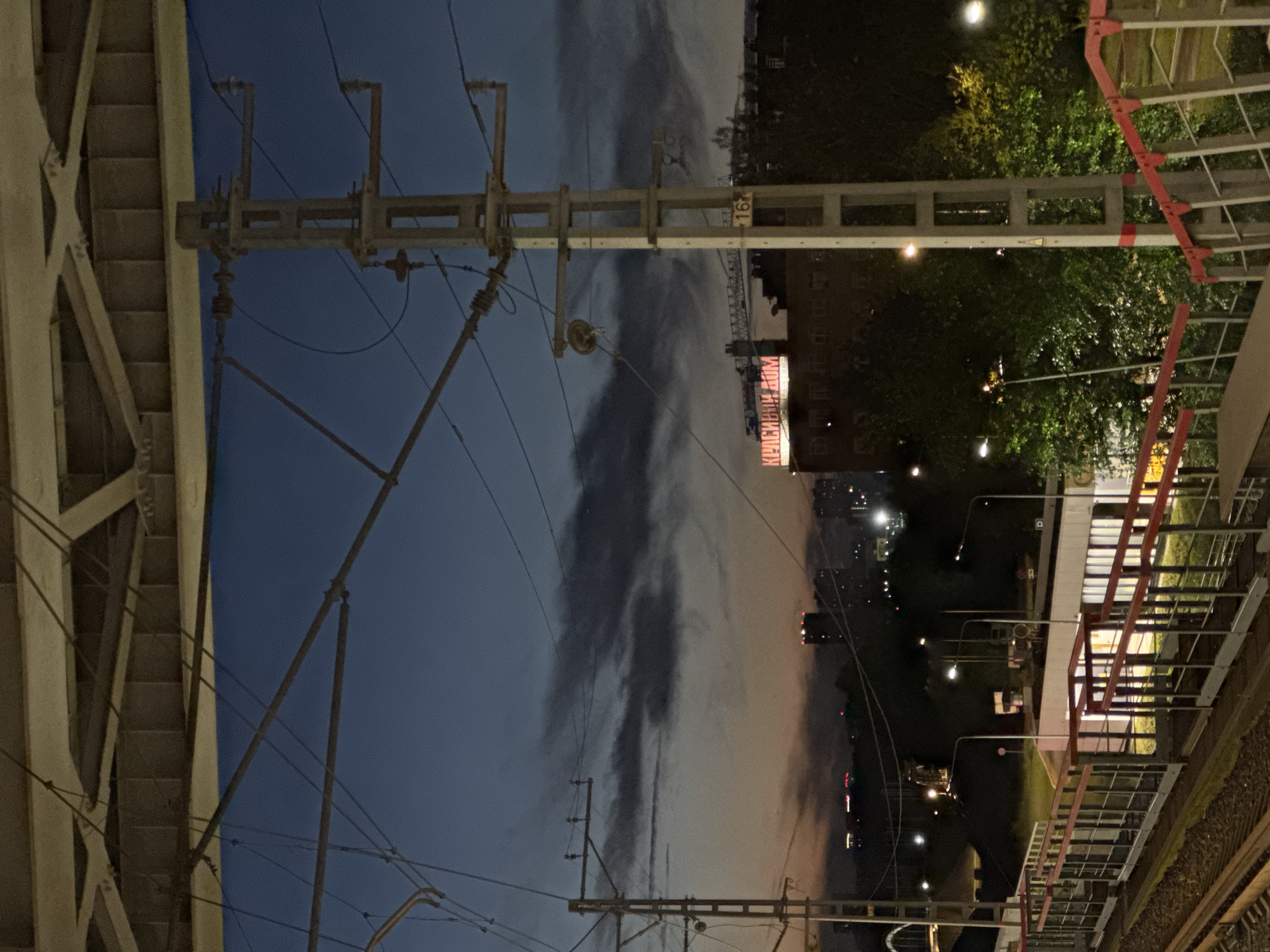 The image depicts an urban scene at dusk, possibly near a train station or elevated railway line, in Moscow, Russia. The sky is a deep blue with wispy dark clouds, transitioning to an orange glow near the horizon. In the background, several tall buildings and city lights are visible, indicating a densely populated area.
Dominating the foreground are overhead power lines and utility poles supporting them, a common sight in areas with electric transportation. A concrete utility pole with multiple insulators and wires is centrally located. To the left, another similar pole and a network of wires crisscross the frame.
Below the power lines, an elevated pedestrian walkway with red railings runs diagonally across the image. The walkway appears to lead down towards a train track, with the rails clearly visible in the lower left corner. A building with lit windows is situated beyond the walkway, and a construction crane is visible above it. A prominent neon sign on a building reads "КРАСИВЫЙ ДОМ" (Krasivyy Dom), which translates to "Beautiful House" in Russian.
The overall atmosphere is one of twilight in a functional urban environment, with infrastructure for transportation and city life illuminated against the darkening sky. There are no people visible in the image.