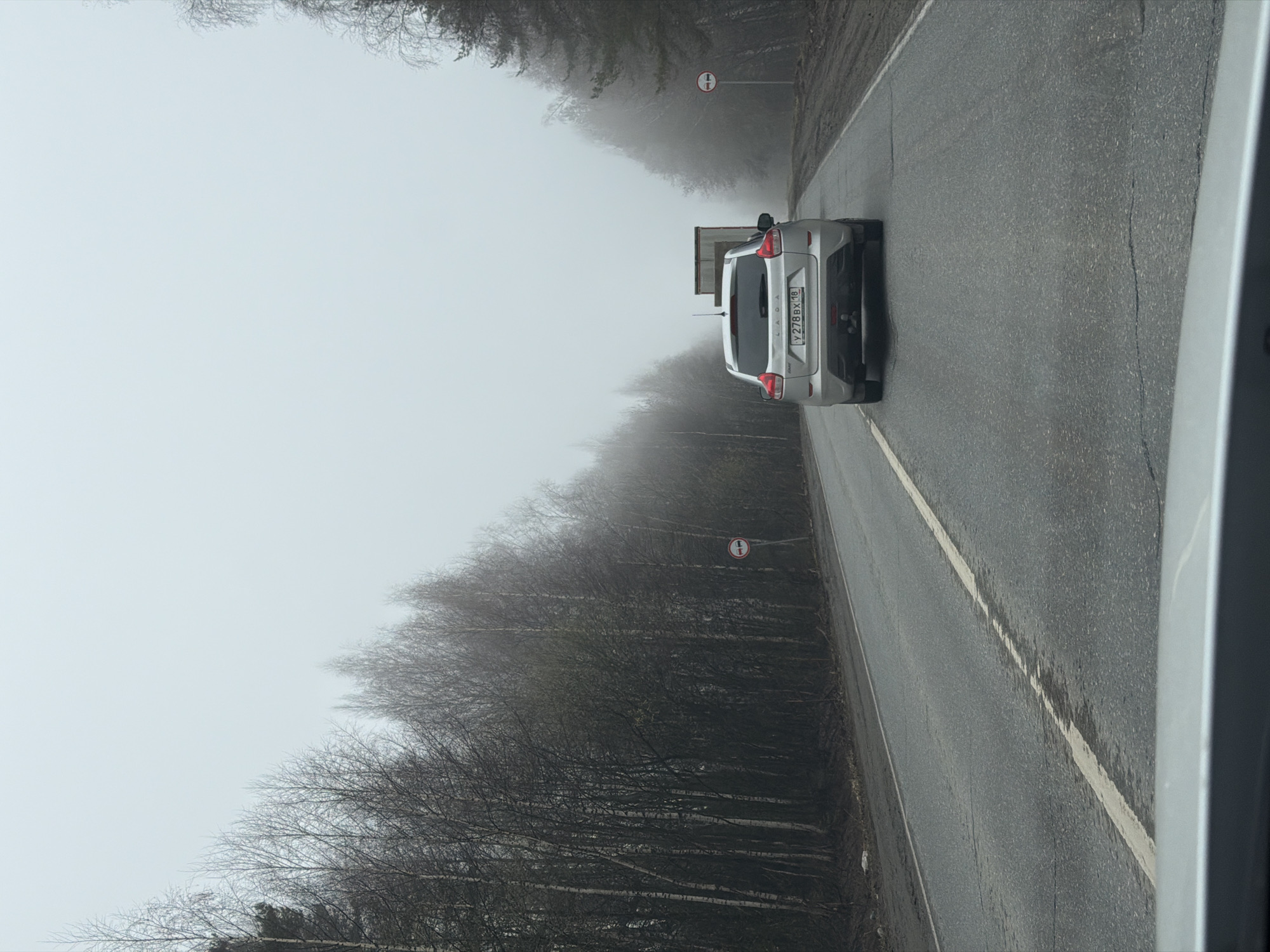 The image captures a misty landscape featuring a light-colored LADA SUV driving away from the viewer on a damp asphalt road. The road stretches into the distance, flanked by dense rows of bare deciduous trees that gradually fade into the heavy fog or atmospheric haze. The sky is uniformly light and overcast, blending almost seamlessly with the low visibility conditions.
On the rear of the SUV, a Russian license plate is clearly visible, displaying the alphanumeric code "Y278BX 18". The brand "LADA" is also visible on the tailgate. Further down the road, on the right side, a red and white circular road sign is posted. The sign depicts two cars (one red on the left, one black on the right) with a diagonal line through them, indicating a "No Overtaking" restriction. The photograph appears to be taken from inside another vehicle, with a portion of its right-side frame visible in the foreground. The overall atmosphere is dim and muted due to the dense fog, suggesting a cool day in late autumn or winter. The location is Bolgury, Russia, as indicated by the region code on the license plate.