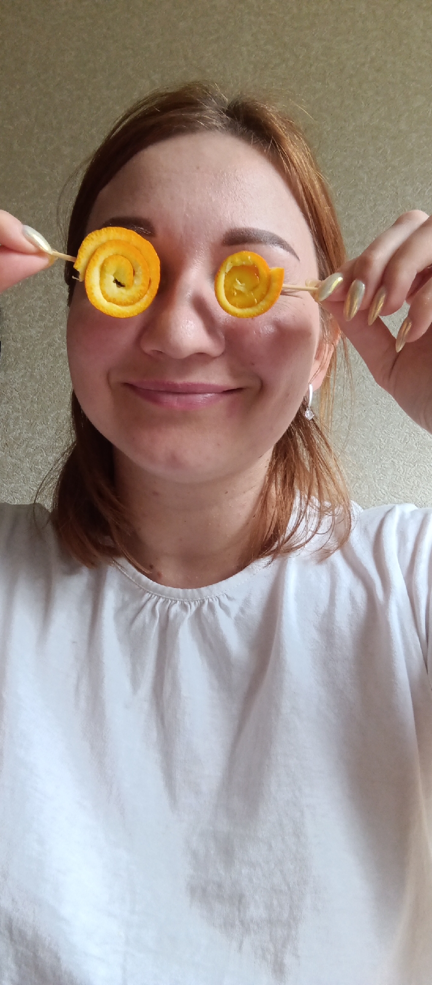 A person is posing with two orange peel spirals held on skewers in front of their eyes. The person has light skin, red hair, and is wearing a white t-shirt and silver earrings. The person is smiling. The background is a beige-colored wall. Location: Unknown, Unknown.
