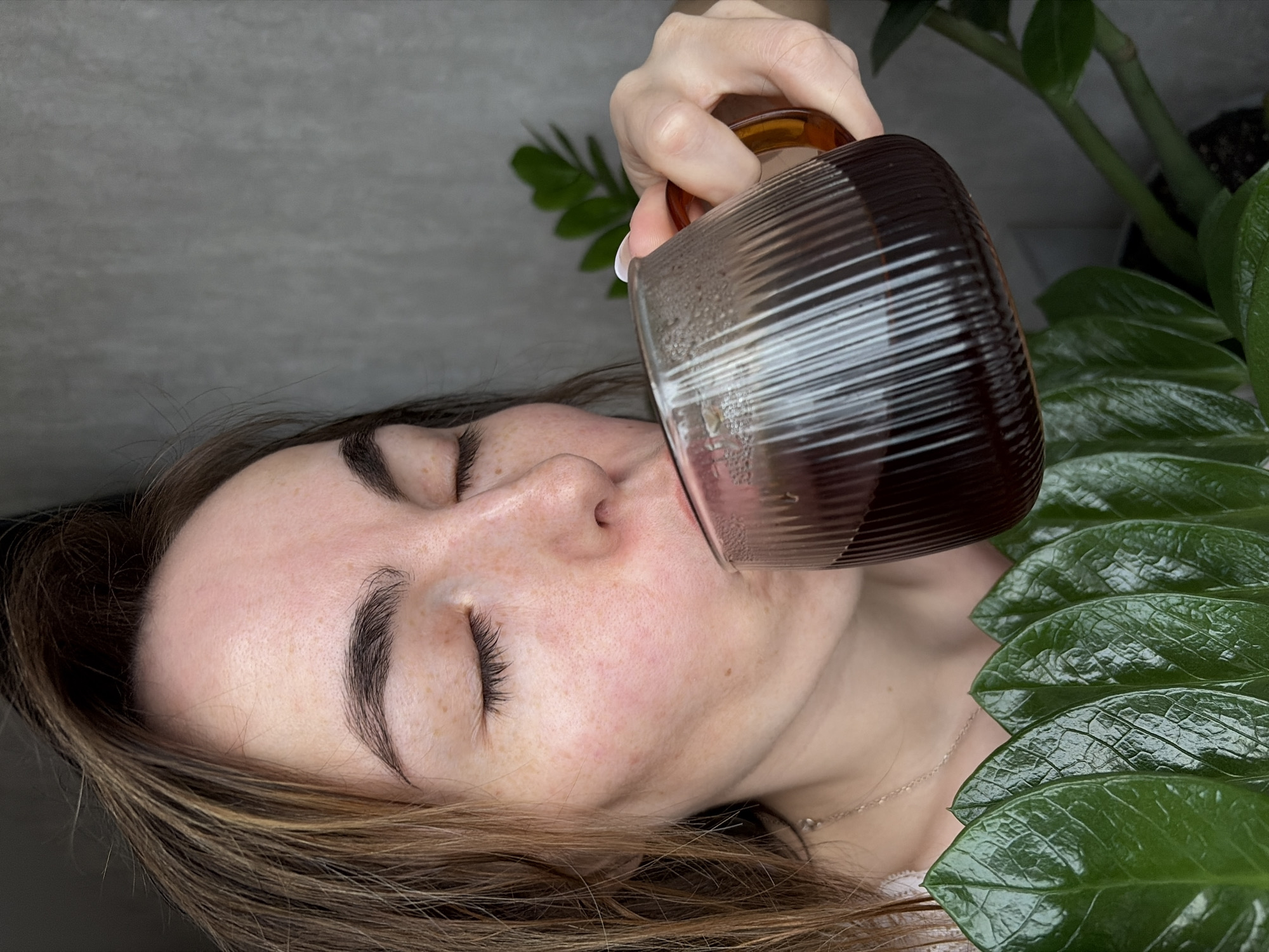 A woman is shown in a close-up shot, tilting a glass of dark liquid toward her mouth. She has her eyes closed, fair skin with freckles, and light brown hair. The glass is ribbed and appears to be filled with a dark-colored beverage. In the background, a potted plant with large, glossy green leaves is visible on the right side of the image, and a neutral gray wall is on the left. The location is not evident from the image.