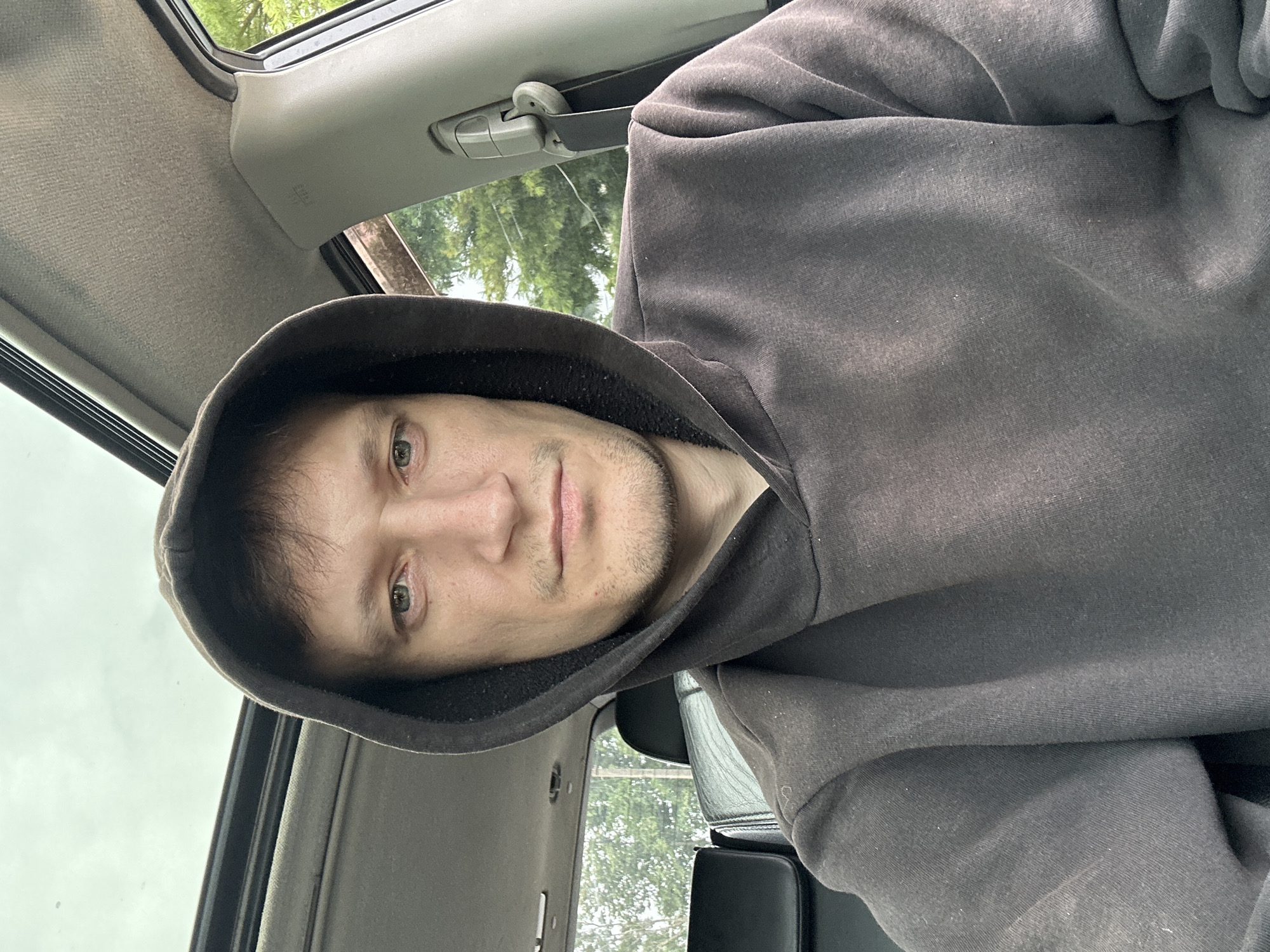 A young Caucasian man with light skin, short brown hair, and a light stubble is taking a selfie in the driver's seat of a car. He is wearing a dark gray hooded sweatshirt, with the hood pulled up. The car's sunroof is open, revealing a cloudy sky. The interior of the car is visible, including the dashboard, a seat, and a seatbelt. Trees and greenery are visible through the open sunroof and the car's windows. The overall setting appears to be an outdoor environment, possibly a street or parking lot in Makiv, Ukraine, based on the provided context.