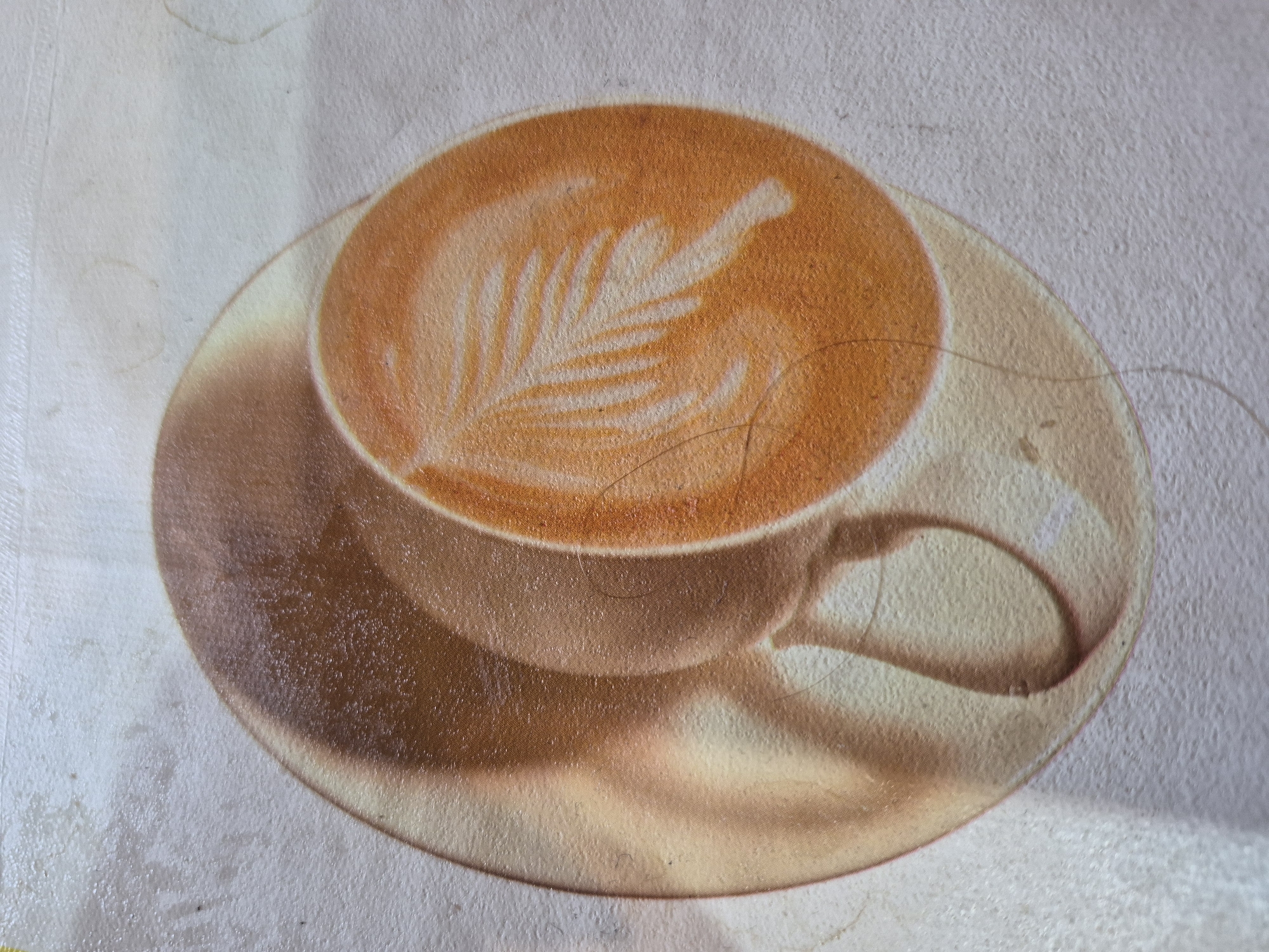 The image is a close-up, overhead view of a latte art on a coffee cup placed on a saucer. The latte art depicts a stylized leaf pattern in white foam on top of an orange-brown coffee. The coffee cup and saucer have a slightly worn, textured appearance, suggesting they might be part of a placemat or a printed surface rather than actual ceramic items. There are faint, circular stains and some loose hairs visible on the surface. The background is a light, textured color, likely a wall or table surface. The image does not contain any specific landmarks or elements that would allow for identification of a location within the city of Yerevan, Armenia.