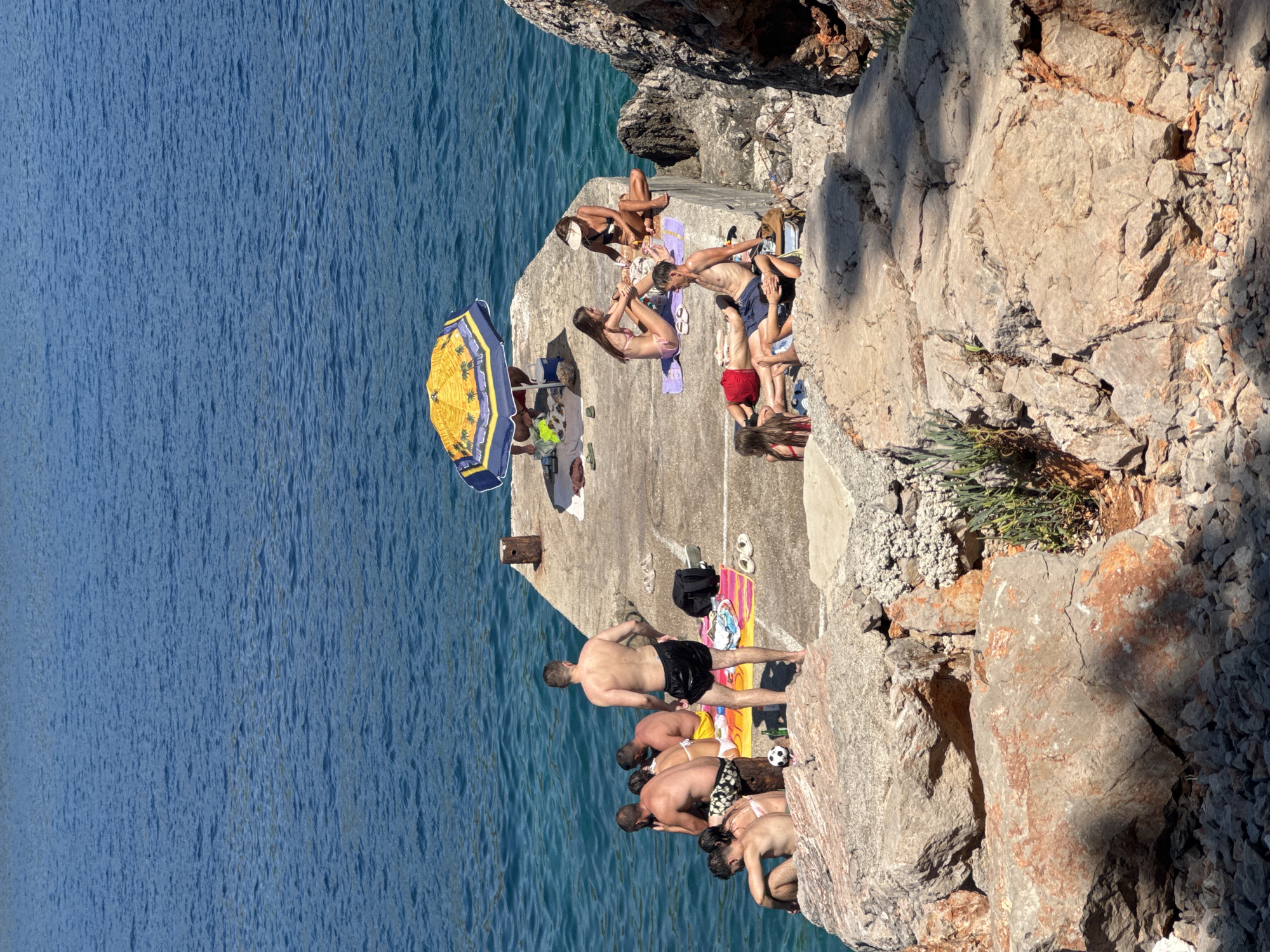 In Dubraljevina, Montenegro, several individuals are observed on a concrete pier and surrounding rocks, engaged in various activities associated with a sunny day by the sea. The water is a vibrant blue, indicating a clear, calm condition. On the pier, a yellow umbrella with a palm tree motif provides shade for a group of people. One individual is seated underneath it, partially obscured. Further along the pier, a woman in a bikini is seen sitting on a towel, facing away from the camera. Next to her, another woman in a bikini is also seated, her posture suggesting relaxation or conversation. A man in dark shorts is lying on his stomach on the pier, with another person beside him also lying down.
To the left of the pier, on a rocky outcrop, a group of individuals are seated, some facing the water and others interacting with each other. One man is standing, looking towards the right side of the image. A football rests on a colorful towel near this group. Scattered on the pier are various objects including towels, flip-flops, a backpack, and a bottle. The environment consists of the concrete pier, large natural rocks, and the expansive blue sea.