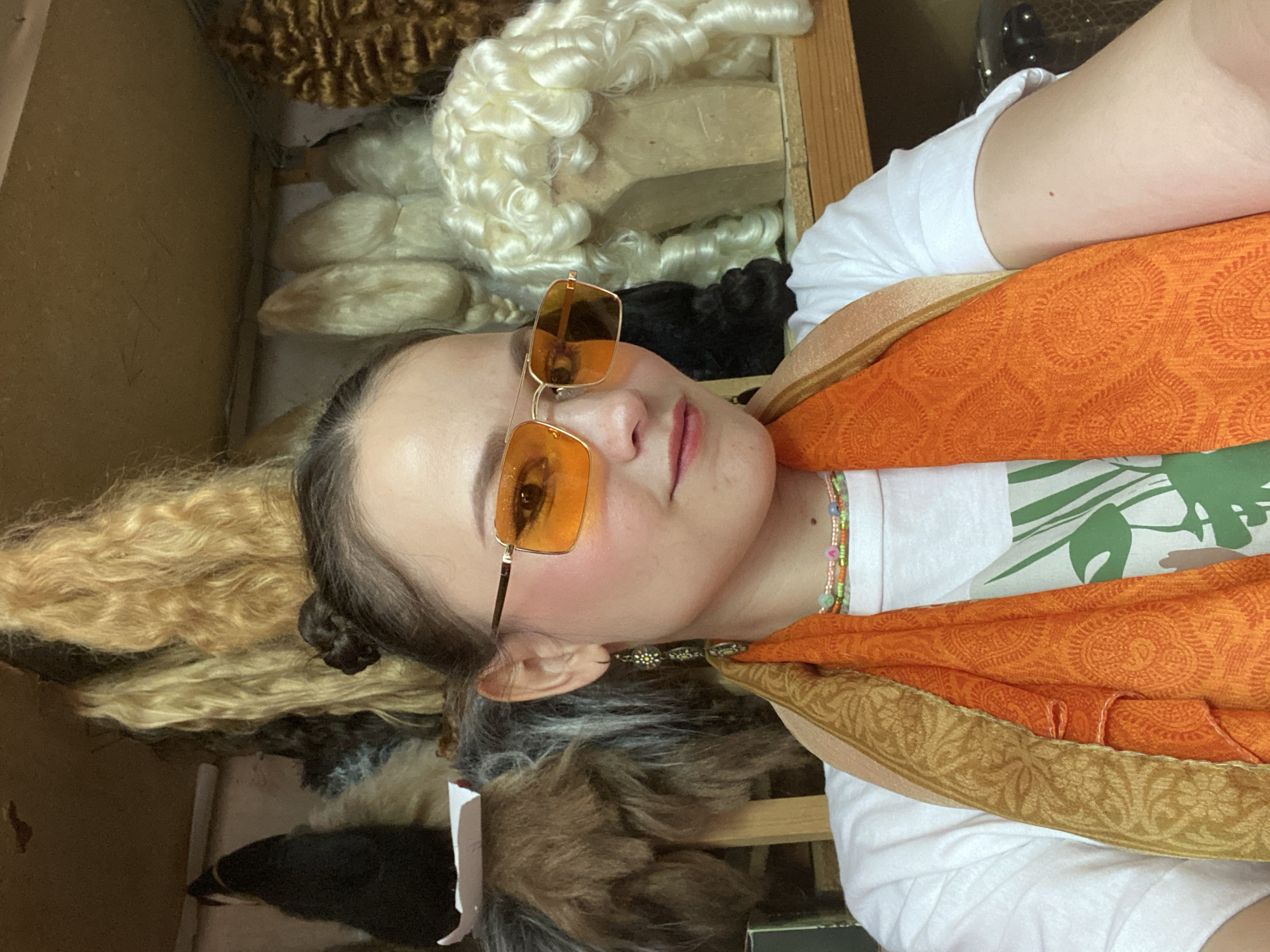 This media file is a portrait of a young person wearing sunglasses and an orange scarf. The subject is looking at the camera with a relaxed expression. The background appears to be a room filled with various wigs, suggesting a wig shop or a costume storage area. There are wigs of different colors and textures visible, including blonde, white, and dark brown. The lighting suggests an indoor setting, possibly during the daytime. The image has a candid feel, as if taken by the subject themselves. There is no visible text in the image. The overall mood is casual and perhaps a bit playful due to the colorful accessories and the unusual setting.