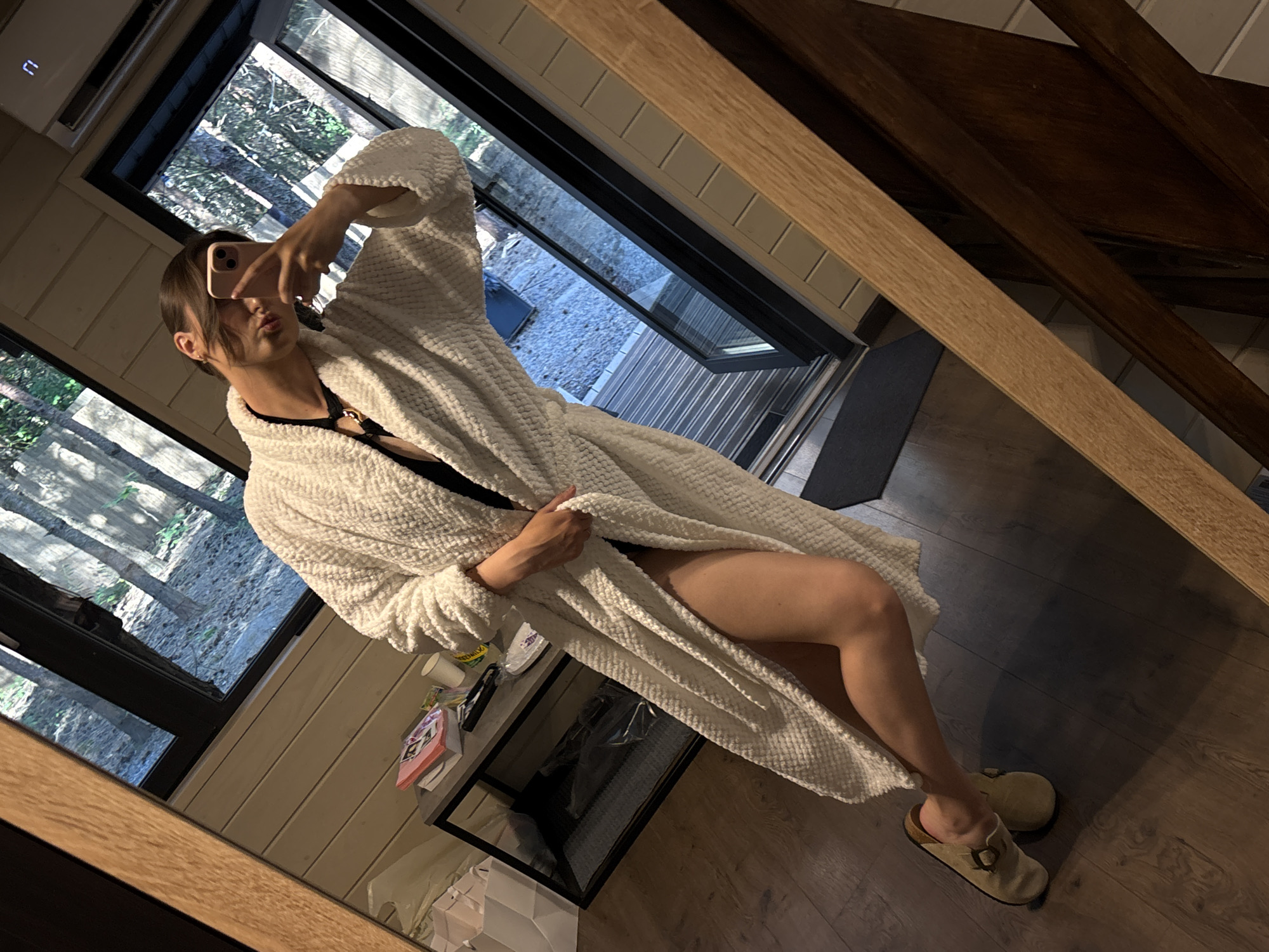 A person, wearing a white bathrobe and tan slip-on shoes, is reflected in a mirror, taking a selfie with a pink smartphone. The person is also wearing a black swimsuit or lingerie. The setting appears to be indoors, possibly a cabin or house, with wooden walls and flooring. Large windows in the background reveal an outdoor scene with trees. On a small table next to the person, there are some papers and a cup. The overall impression is a casual, personal moment captured by the individual.