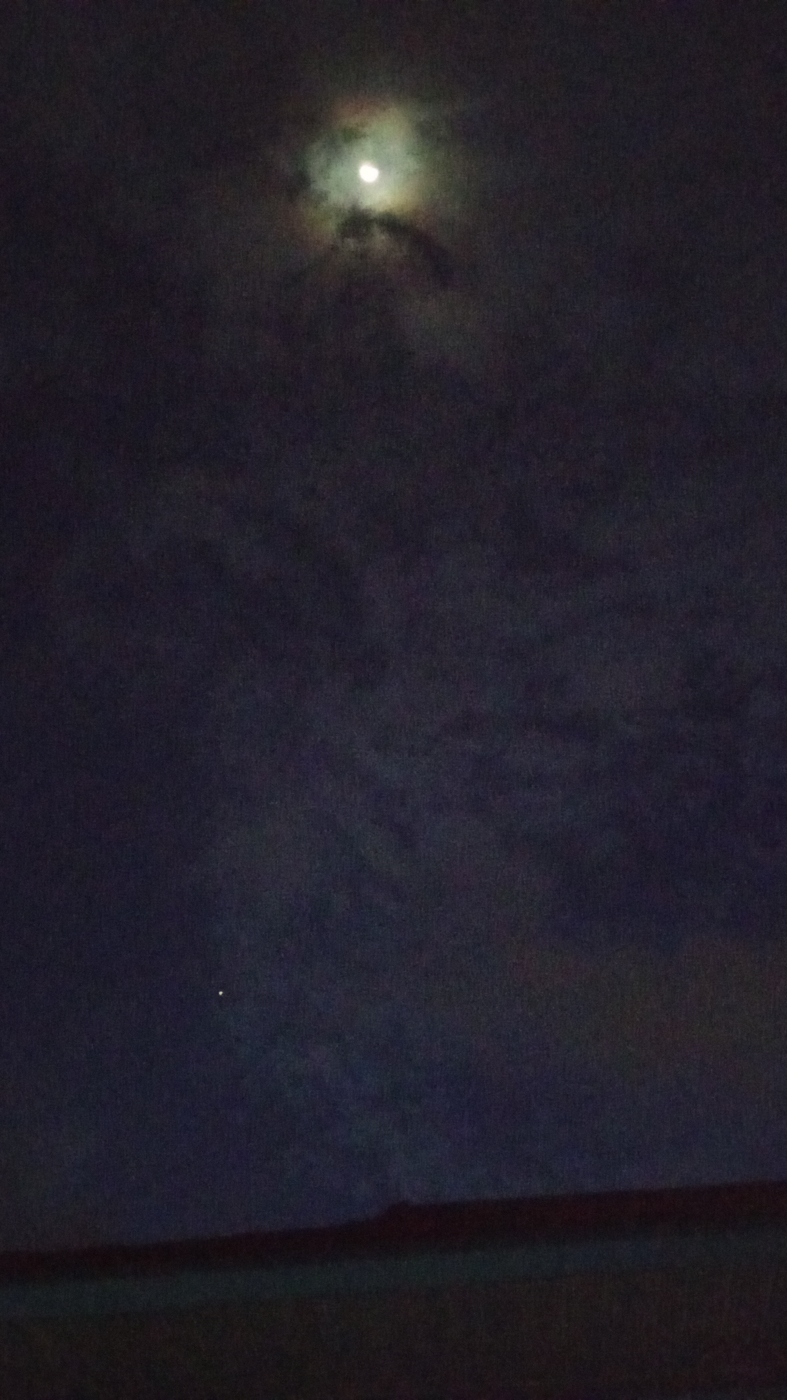 The image shows a night sky in Lhokseumawe, Indonesia. A partially obscured moon is visible near the top of the frame, surrounded by clouds. A single bright star is visible in the lower left portion of the sky. The bottom of the image reveals a dark, indistinct horizon line, possibly representing buildings or vegetation. No people or interactions are discernible. The environment is dark and cloudy.